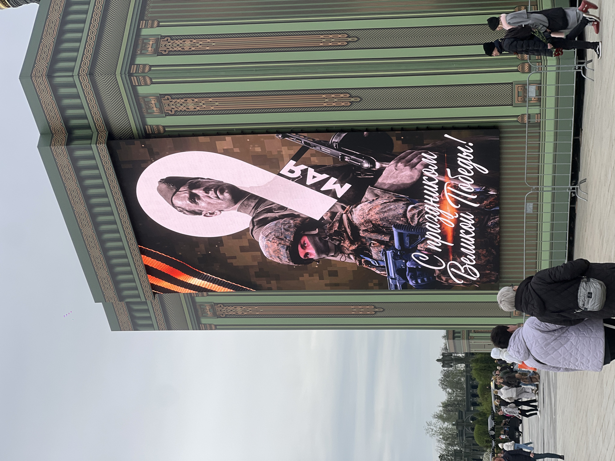 The image depicts an outdoor scene in Zelenaya Roshcha, Russia. A large LED screen displays an image of a soldier holding a firearm. An orange and black ribbon is visible, along with the words "С праздником Великой Победы!" which translates to "Happy Great Victory Day!". The image also includes the word "МАЯ", meaning "May".
Several people are walking in the foreground, and a metal fence separates the screen from the crowd. The screen is mounted on a green and tan patterned structure. The sky is overcast.