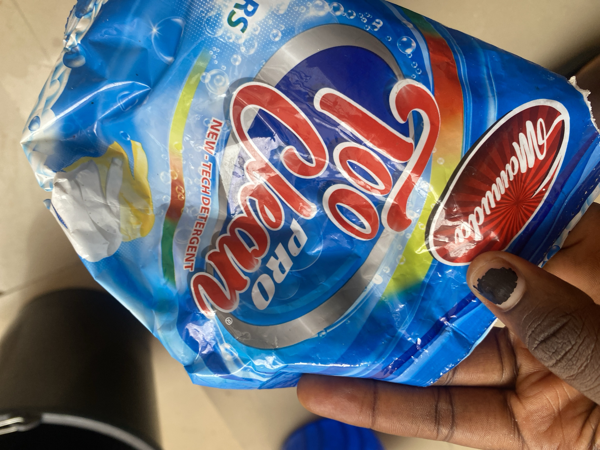 A hand is holding a crumpled blue plastic bag of "Pro Lean" detergent. The bag has a vibrant design with red lettering and yellow and green stripes. There is a circular logo on the right side with the word "Mamuda" in white script. The bag appears to be in a domestic setting, possibly a home in Bulamare, Nigeria, given the provided location context. The hand holding the bag has dark skin and a black painted fingernail. The lighting suggests it could be daytime. There are no other people or significant environmental details visible in the image.