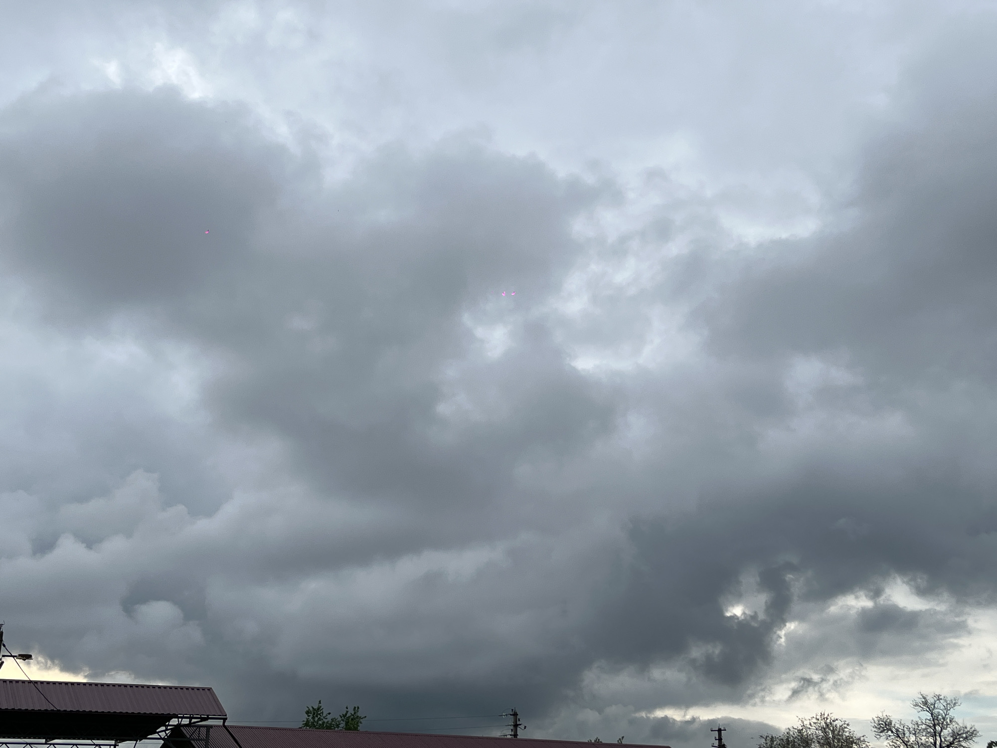 The image shows a cloudy, overcast sky in Novopokrovskaya, Russia. The sky is filled with dark gray clouds, suggesting an impending storm. In the foreground, the roofs of buildings are visible, with a dark brown color. Power lines and trees are also seen at the bottom of the frame. There are a few faint pink spots visible among the clouds.