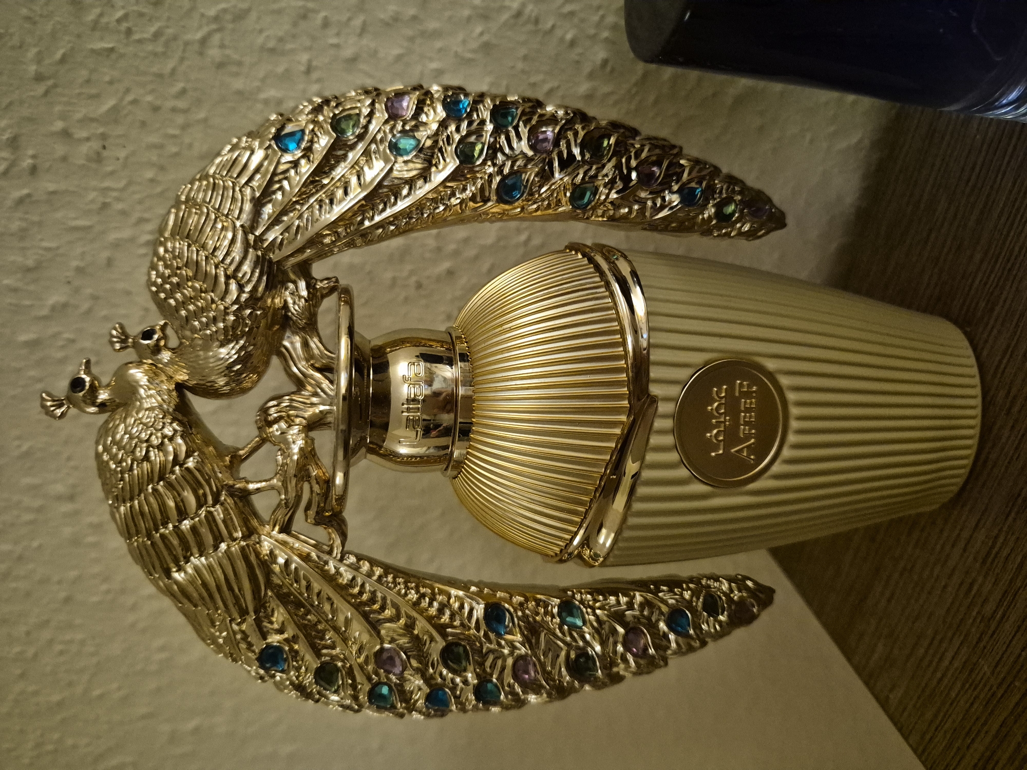 A gilded perfume bottle sits on a wooden surface. The bottle is fluted and has a gold cap with two decorative peacocks perched on top. The peacocks have colorful jewel-like accents on their wings. The brand name "Lattafa" is visible on the cap, and "Areef" is written in Arabic and English on a round medallion on the bottle's body. The background is a textured, light-colored wall.