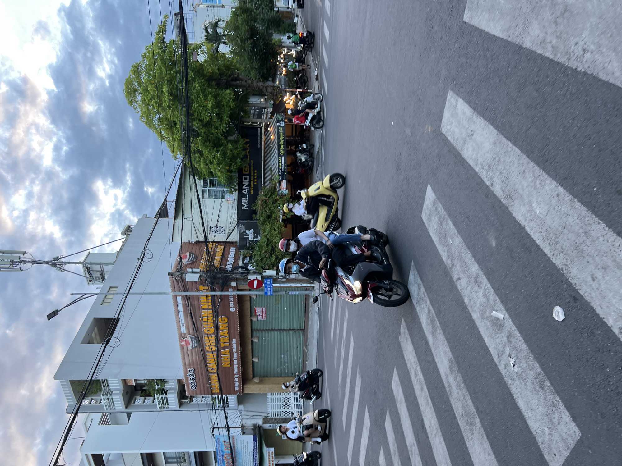 In Nha Trang, Vietnam, a busy intersection is captured under a cloudy sky. The foreground features a pedestrian crosswalk on an asphalt road. Several individuals on motorcycles are navigating the intersection. In the mid-ground, two people are riding a red and black motorcycle, the driver wearing a blue helmet and a dark jacket, and the passenger wearing a white shirt and a red helmet. To their left, a woman on a beige scooter wears a white shirt and a white helmet. Further back, a person on a dark scooter wears a white helmet.
The background is lined with buildings, including a white building with balconies and greenery, and a brown building with signage for "BÁNH CANH CHE QUAN NHAT". Another building displays "MILANO GALAXY" and "CAFE SANG TRONG, HIEN DAI, CHAT". Streetlights and numerous overhead wires crisscross the scene. A tall tower with multiple antennae stands prominently against the sky. The overall atmosphere suggests a typical urban street scene in Vietnam.