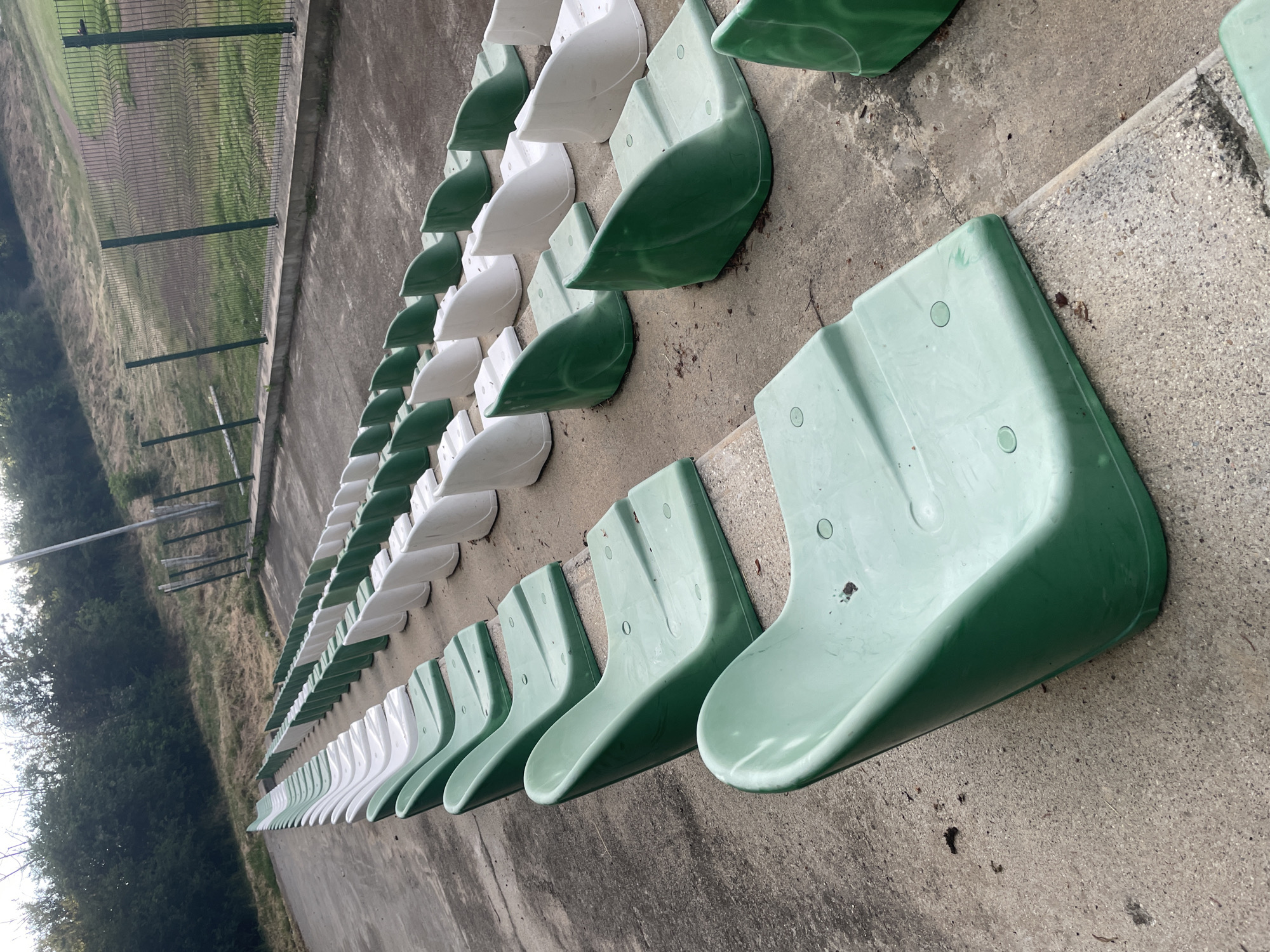 The image captures a long row of empty stadium seats at an outdoor sports facility, likely a small stadium or pitch. The seats are made of molded plastic and alternate in color between a muted green and white, creating a pattern along the concrete bleachers. The perspective is from a low angle, looking upwards and along the length of the seating area.
To the left of the seats, a green wire mesh fence runs parallel, separating the concrete seating from a sloped, grassy field that appears dry and somewhat unkempt. Beyond the field, a line of trees is visible under what appears to be an overcast sky or diffused daylight, as strong shadows are absent. The concrete surfaces around the seats show signs of weathering and some scattered debris.
No people are present, indicating that no event is currently underway. The scene conveys a sense of quiet emptiness, perhaps awaiting spectators. There is no discernible text or signage visible in the image. Based on the provided context, the location is Ihtiman, Bulgaria.