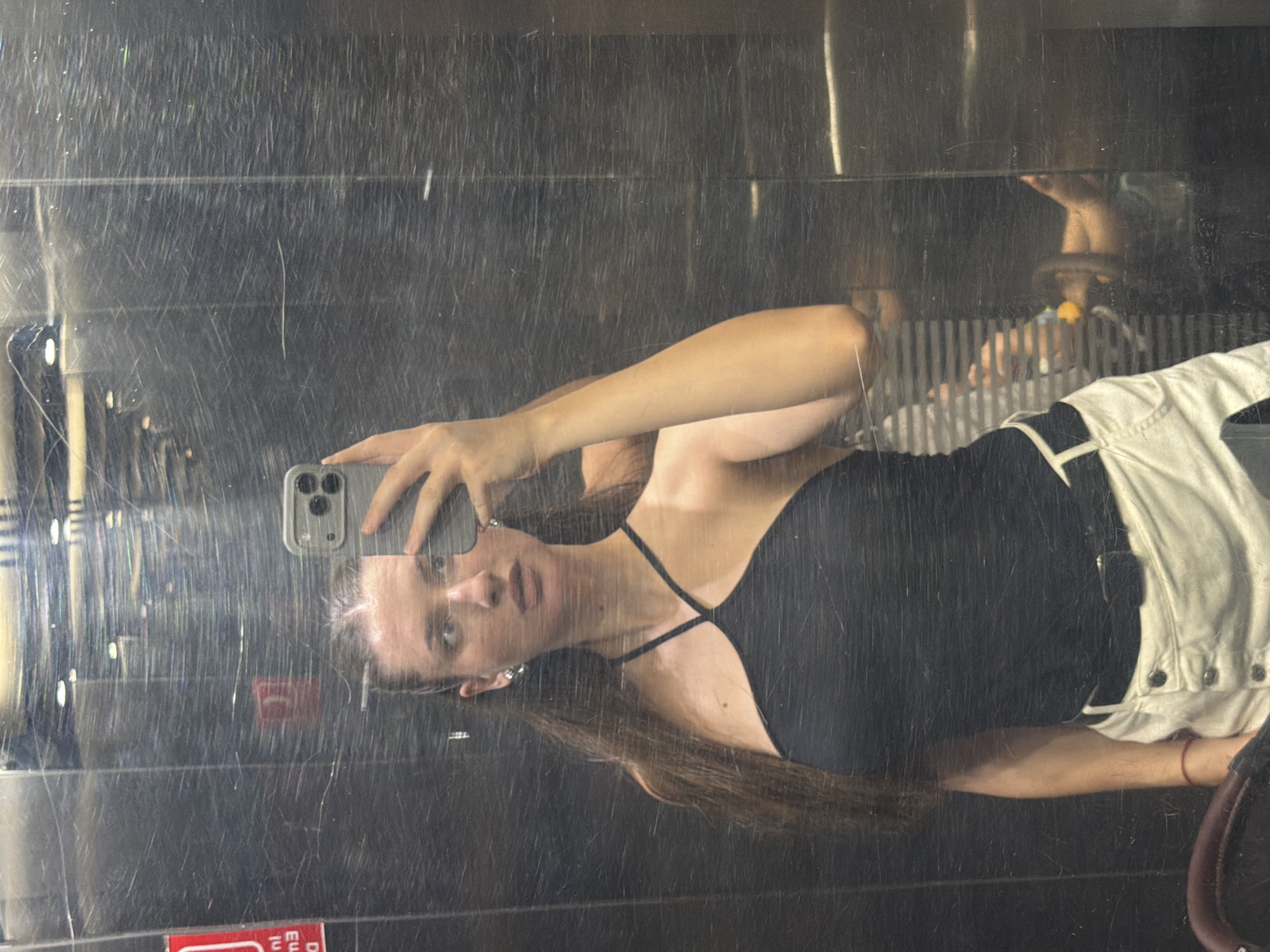 A young woman with long, dark brown hair pulled back into a ponytail is taking a selfie in a scratched, metallic elevator. She is wearing a black halter top and white shorts with a black belt. Her left arm is raised, holding a gray smartphone, and her right arm is bent at the elbow, resting on her hip. Her eyes are wide and looking towards the camera. The elevator walls are heavily scratched, reflecting lights from above and the surrounding environment. A red sign with white lettering is visible on the left wall. In the reflection, a person's legs can be seen in the background, suggesting another person is in the elevator. The elevator appears to be in a public building, potentially a hotel or resort in Sharm El Sheikh, Egypt, given the context.