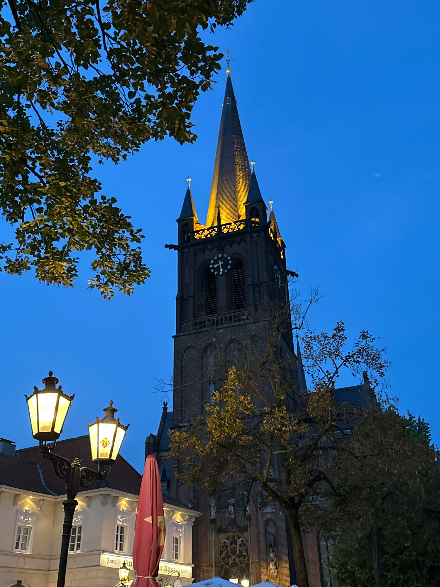 This is a vertical, outdoor, eye-level shot of the exterior of the Lutherkirche in Krefeld, Germany, at dusk. The church tower, with its tall spire lit from below, dominates the right side of the frame. Gargoyles protrude from the tower, and a clock face is visible on its side. The sky is a deep, twilight blue. In the foreground, to the left, are two ornate street lamps casting a warm glow. Behind the lamps is a white building with traditional European architecture, featuring multiple windows and decorative elements. A closed red umbrella stands prominently in the mid-ground, and trees with sparse, autumnal foliage are scattered throughout the scene, partially obscuring the church and buildings. The image appears to capture an evening ambiance in a European city square.