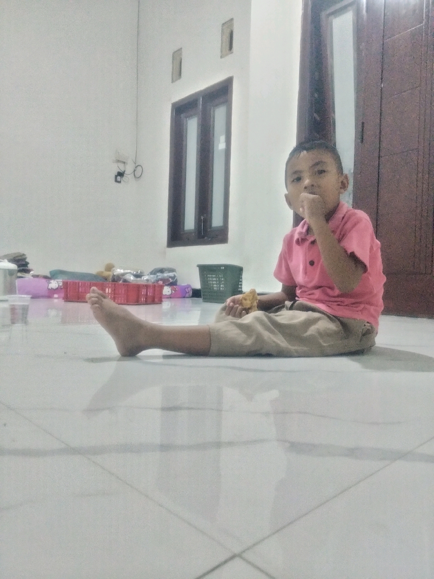 A young boy wearing a pink polo shirt and tan pants sits on a tiled floor, eating. He is positioned in the center of the frame, with his legs extended towards the viewer. In the background, a window with dark panes is visible on a light-colored wall. To the right of the window, a wooden door is present. Various objects, including a green basket and a red plastic crate filled with colorful items, are scattered around the floor in the midground. The overall environment appears to be an indoor space.