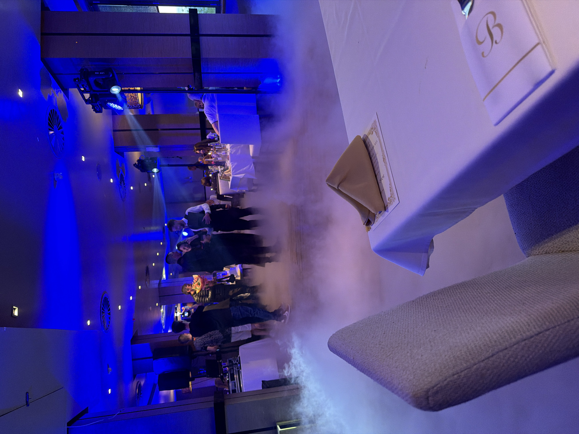 Here is a detailed description of the image:
**General Overview:** The image appears to be taken inside a large, modern banquet hall or event space. The room is lit with blue and white lighting, creating a dramatic ambiance. There is a mist or fog effect on the floor. The focus is on a table setting in the foreground, while in the background, there is a crowd of people.
**Foreground Details:**
* A white tablecloth covers the table.
* A folded napkin with a gold-colored letter "B" is present on the table.
* There is a place card with gold trim next to the napkin.
* Part of a chair upholstered with a light-colored fabric is visible.
**Background Details:**
* A group of people is standing in the background, possibly socializing or watching an event.
* Some of the individuals are dressed in formal attire.
* There is a DJ booth or stage area with equipment present.
* The walls are decorated with vertical accents and ventilation grilles.
* Overhead lighting fixtures are visible, contributing to the room's lighting scheme.
**Atmosphere:** The presence of fog or mist on the floor suggests a celebration or performance, perhaps a wedding or a formal dance. The blue lighting adds to the festive and somewhat surreal atmosphere.
**Possible Location:**
Given the event's scale, the type of architecture, and the presence of signage in Bulgarian, it's probable the event is in Sofia, Bulgaria, although there is no way to confirm based solely on the given picture.