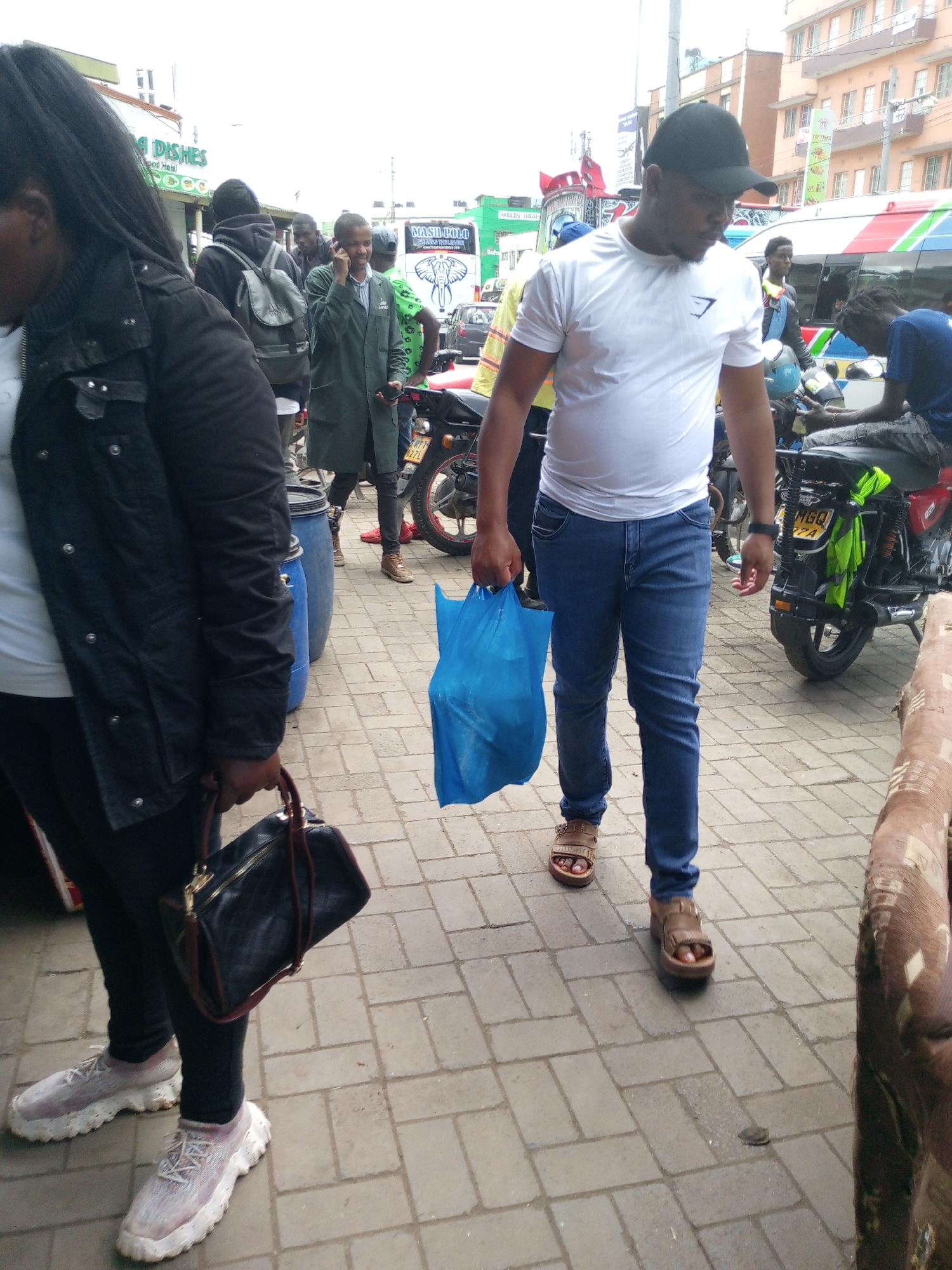 The image depicts a bustling street scene in Nairobi, Kenya. The ground is paved with interlocking concrete blocks.
A woman on the left, partially in the frame, wears a black jacket, black pants, and white sneakers. She carries a black handbag. Next to her is a man wearing a white t-shirt, blue jeans, and brown sandals. He holds a blue plastic bag. In the background, several people are walking or standing around. One man wears a green coat and is on a phone call. Several motorcycles are parked in the background, along with a public transportation vehicle. Signage for "MASH PO LO" and "KENYA DISHES" is visible.