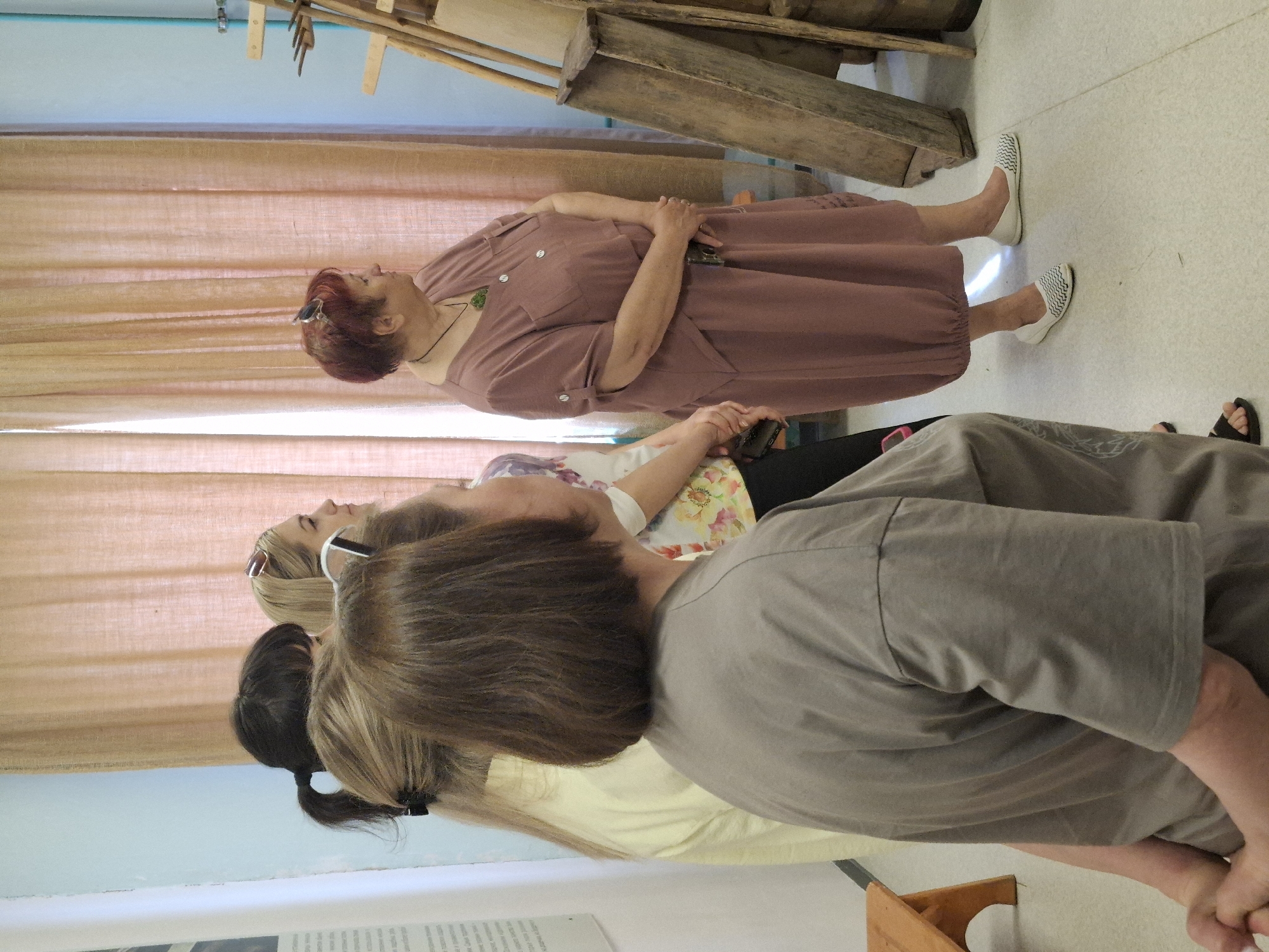 The image shows a group of women indoors, likely in the city of Markovskii, Russia. One woman, with red hair and wearing a brown dress, stands with her hands clasped in front of a curtain backdrop. Several other women are present, one wearing a grey shirt and facing away from the camera, and another with blonde hair and sunglasses. In the background are wooden objects, possibly tools or furniture, suggesting a museum or workshop environment. The floor is tiled in white.