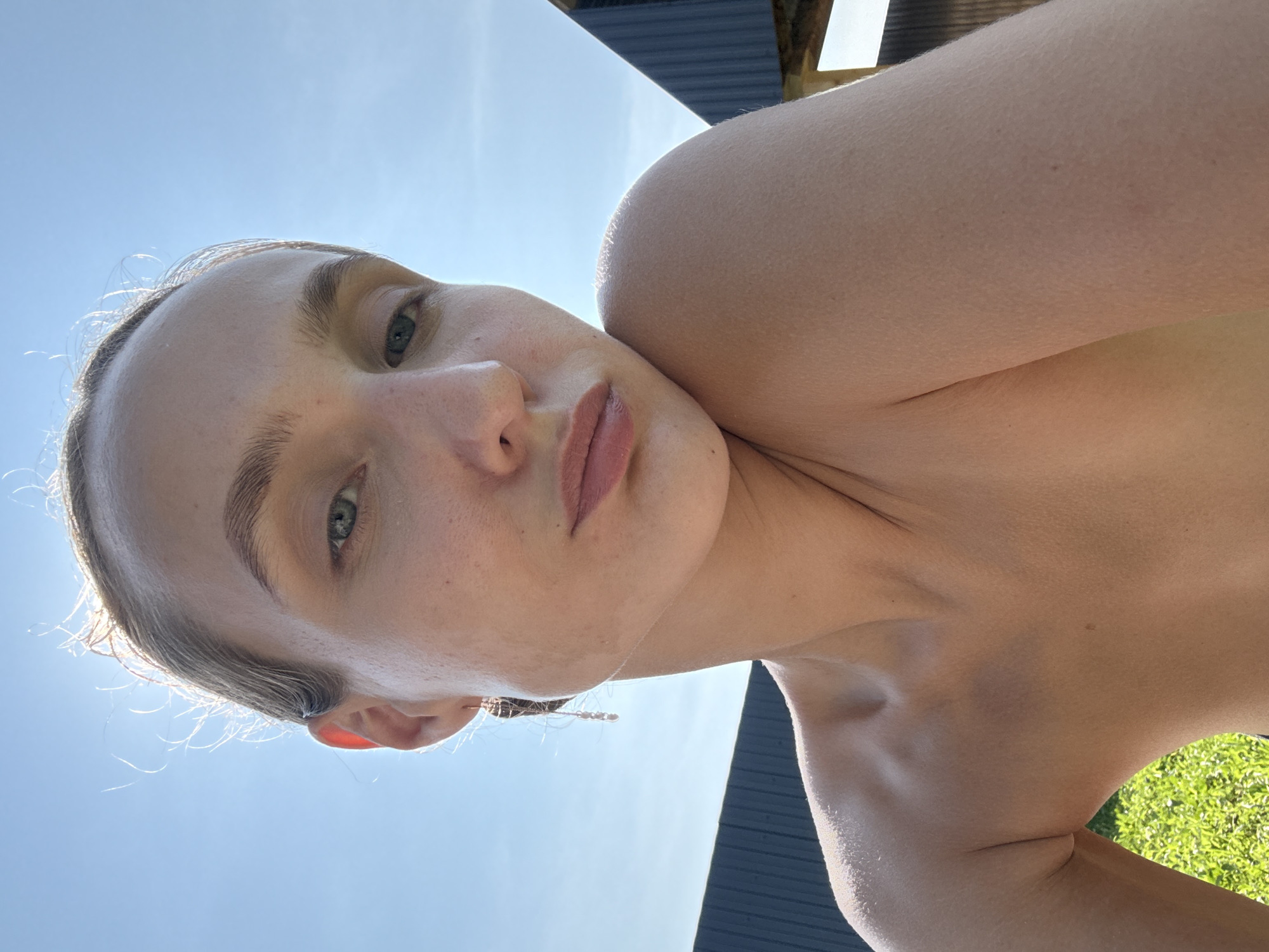 A young woman is captured in a close-up, low-angle selfie. She is lying down, looking upwards towards the camera. Her face is illuminated by the bright sunlight, creating a soft glow and highlighting her features. Her eyes are open and looking directly at the viewer. Her lips are slightly parted. Her hair is pulled back, with some wisps of hair catching the light around her forehead and ears. She appears to be outdoors, with a clear blue sky visible above. In the background, there is a dark, textured surface, possibly a roof or a fence, and a patch of green grass. The overall impression is one of a relaxed and candid moment captured on a sunny day. The time of day is likely daytime, given the strong sunlight. The location context provided is Подшивалово, Russia. There is no visible text in the image.