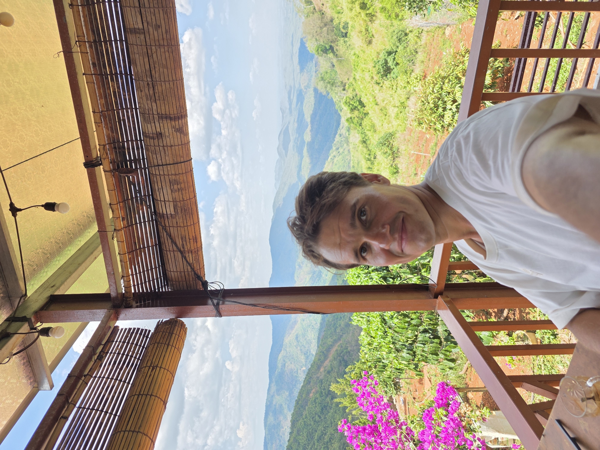 A medium-shot captures a person with short brown hair and fair skin, wearing a white t-shirt, partially visible in the frame. The individual is smiling, facing slightly towards the camera. The background features a panoramic landscape of lush green hills and valleys under a partially cloudy sky. A structure with a bamboo ceiling and a wooden railing frame the view, suggesting the setting is a balcony or terrace. In the foreground, vibrant pink flowers add a pop of color against the green foliage. The location is Nao Sẻ, Vietnam.
