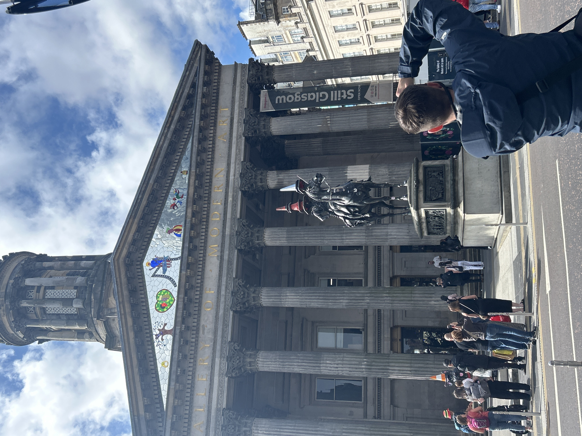 The image shows the exterior of the Gallery of Modern Art in Glasgow, Scotland. The building is grand and classical, with a dome at the top and tall columns. A statue of a person on horseback is in front of the gallery. The statue is wearing conical hats. Several people are standing on the sidewalk in front of the gallery, some of whom appear to be tourists. One person in the foreground is taking a picture of the gallery. The sky is blue with some clouds. A banner on the side of the building reads "Still Glasgow". The scene is bustling with activity, suggesting a public space with visitors.