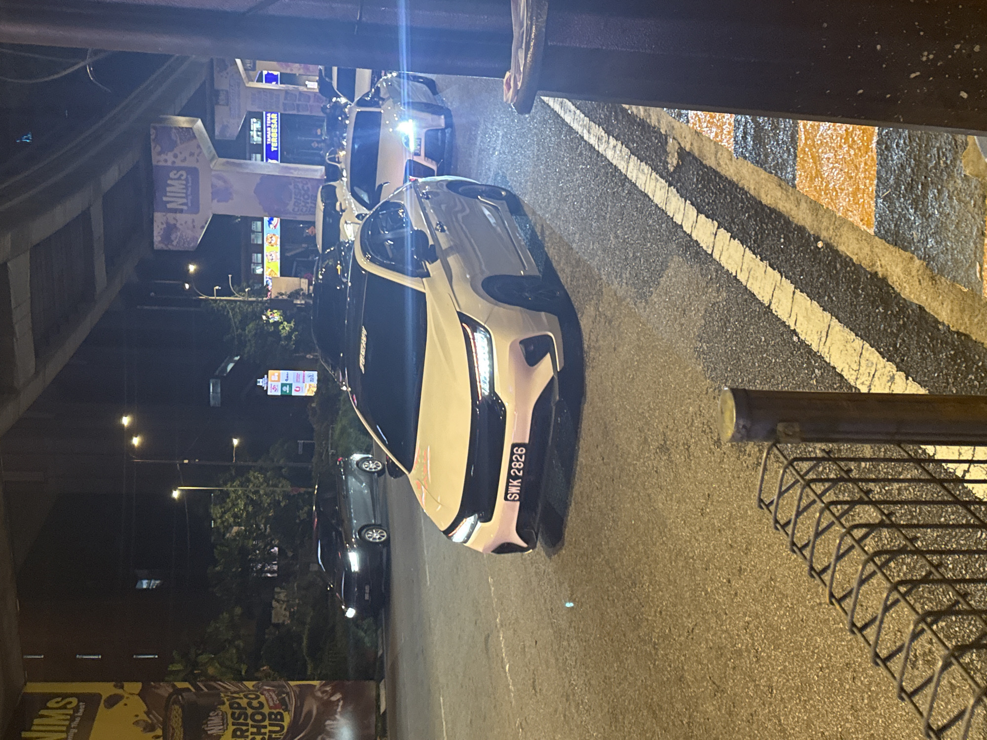 The image captures a nighttime street scene in what appears to be an urban area, likely under an elevated structure or overpass. The camera is positioned looking down at an angle onto a roadway.
The main subjects are several cars. In the foreground, a white or light-colored modern sedan (possibly a Honda City or similar model) is prominent, displaying a front license plate that reads "SWK 2826". Its headlights are on, casting light forward. Another similar white sedan is visible further behind it, also with its lights on. In the background, several other dark-colored cars are visible, some with their lights on, suggesting traffic or parked vehicles.
The road surface is asphalt, marked with distinct white and yellow lines, typical of traffic lanes and perhaps a pedestrian crossing area. A metal barrier or cage-like structure is visible on the right side of the frame, close to the camera's viewpoint.
On the left side, various commercial signs are visible against the dark sky. A large, prominent sign features the word "NIMS" with "CRISPY CHOCO TUB" and smaller text beneath it, likely advertising food products. Another sign further back shows "NIMS" again, along with "TERBESAR" (Malay for 'largest'). Streetlights illuminate parts of the background, revealing some trees and what seem to be commercial buildings. The overall setting indicates a bustling urban environment after dark. The license plate "SWK" suggests the car is registered in Sarawak, Malaysia, consistent with the provided location context of Kuala Lumpur, Malaysia.
