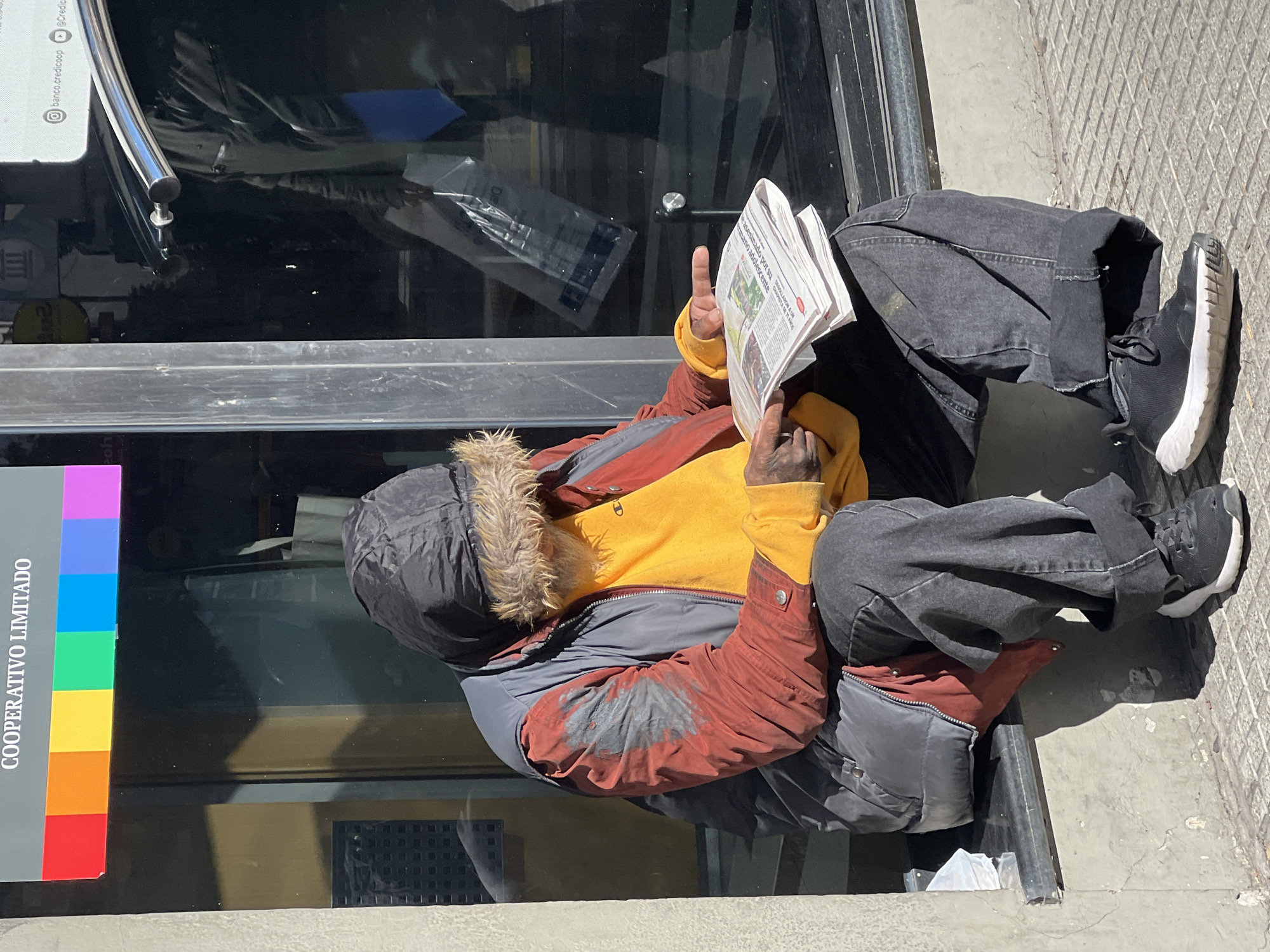 A man sits on the sidewalk next to a building in Buenos Aires, Argentina. He is reading a newspaper. The man is wearing a black beanie with fur trim, a yellow sweatshirt, a red and gray jacket, and black jeans. His shoes are black with white soles. The building next to him has a sign that reads "COOPERATIVO LIMITADO" with various colors of the rainbow.
