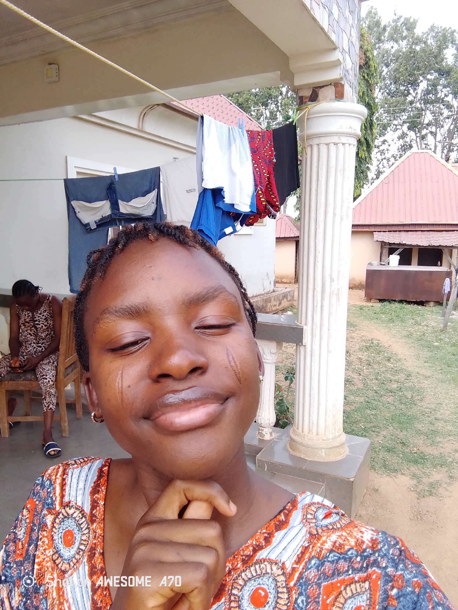 A young woman is taking a selfie on a porch in Unknown, Unknown. The background includes a clothesline with clothes hanging on it, a white pillar, another woman sitting in a chair, a building with a red roof, and grassy areas. The selfie taker is smiling with her eyes closed and has a hand under her chin. She is wearing a patterned shirt.
