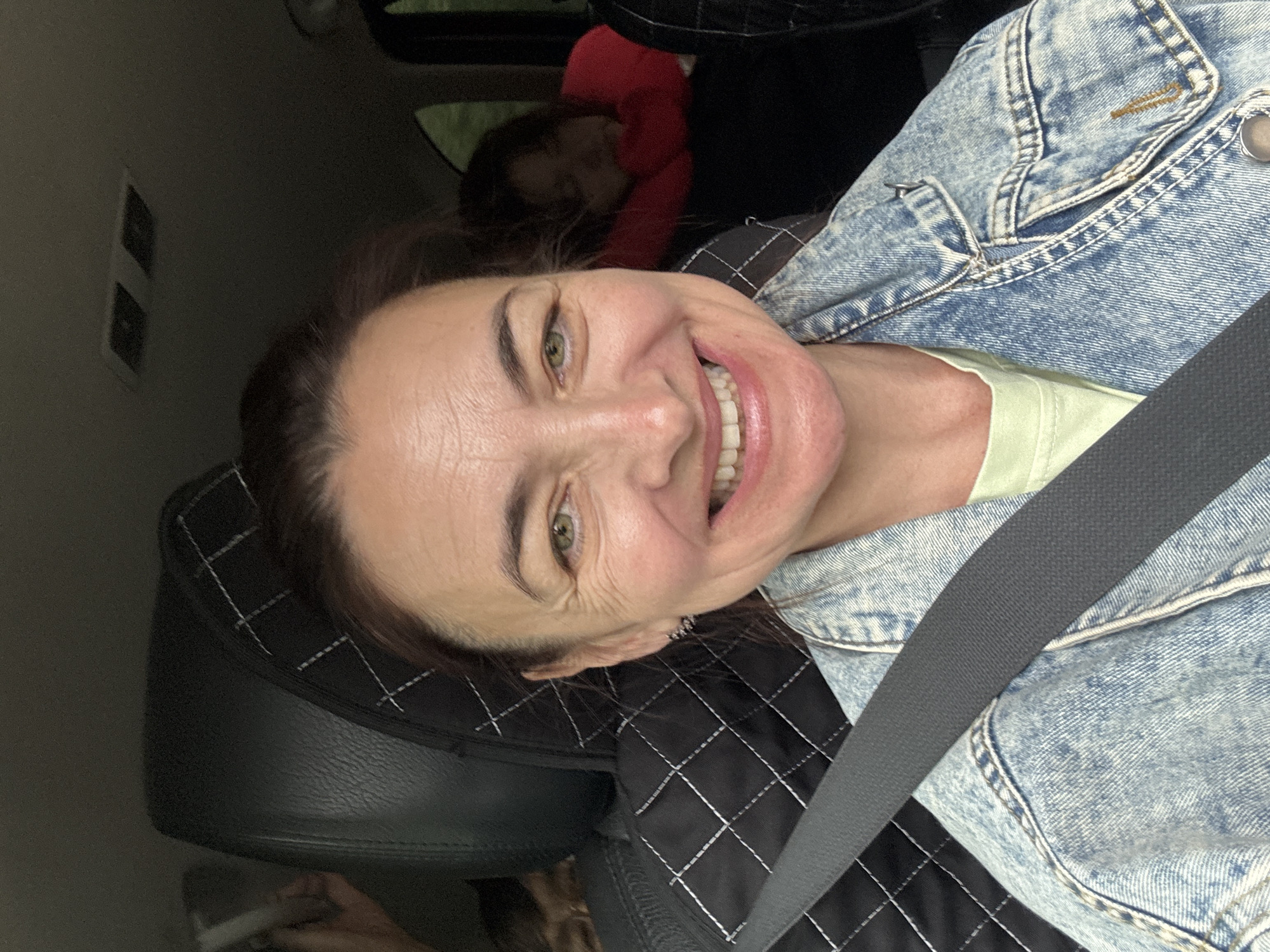 The image shows a woman in the front seat of a car, smiling at the camera. She is wearing a denim jacket over a light green t-shirt and her seatbelt is visible. In the background, two children are seated in the back of the car. The child on the right is leaning on a red pillow and appears to be asleep. The environment is indoors, inside a vehicle, with a dark interior. The image does not provide any specific location details within the city of Kulmas, Russia.