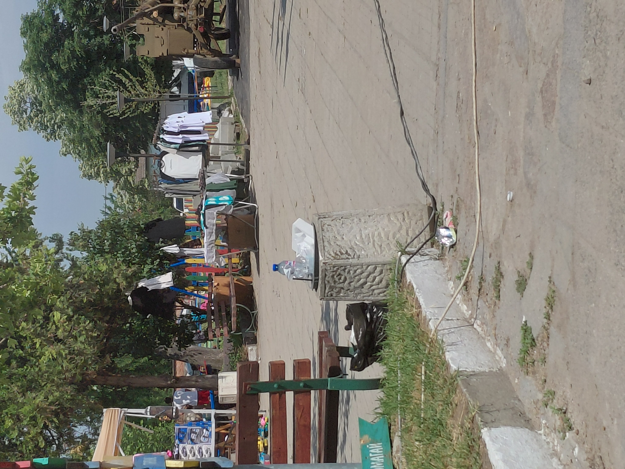 This image depicts an outdoor public area in Kostievo, Bulgaria, consistent with a temporary market or fairground under clear weather conditions.
In the immediate foreground, a patch of grass borders a light grey paved surface. A white-painted concrete curb separates these areas. Running across the pavement are multiple dark electrical cables and one lighter beige cable. Scattered litter, including a small white wrapper and metallic foil, is visible on the ground.
On the left side of the foreground, a wooden park bench with dark green metal supports is partially visible. Adjacent to it, a small, partially visible green sign with white Cyrillic text is present. To the right of the bench, a black plastic bag containing refuse rests on the ground.
In the mid-ground, a square, light-colored concrete or stone pedestal with a textured, possibly carved, surface stands prominently. On its flat top, a transparent plastic water bottle with a blue cap rests next to a white plastic tray holding several white plastic objects, likely cups or plates.
Further into the mid-ground, a series of outdoor market stalls are set up. One stall on the far left features a beige canopy and displays various items, including packaged goods on tiered shelving. Two individuals are partially visible near this stall; one person wears a white t-shirt and red shorts, facing away, while another person's upper body is visible further back. Multiple clothing racks display a variety of garments, predominantly white t-shirts, across several market stands.
Two tall streetlights with dark, disc-shaped luminaires are positioned between the market stalls and the background. Behind some of the market displays, a section of a colorful wooden picket fence, featuring red, blue, yellow, and green vertical planks, is visible. Another wooden bench is situated behind this fence.
The background is dominated by lush green deciduous trees, indicating a park or heavily vegetated area. Above the trees, a light blue sky with minimal cloud cover is observed. On the far right of the image, a large, olive-drab military vehicle, possibly an artillery piece, is static. It features large wheels and a prominent barrel or cannon pointing upwards and to the right.