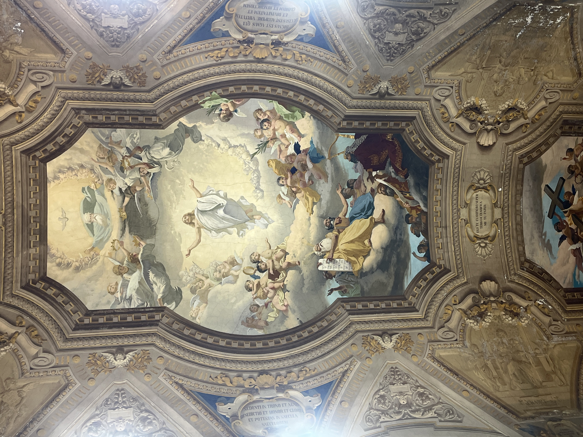 The image depicts a highly ornate ceiling with a large fresco, likely within a church or palace in Buenos Aires, Argentina. The fresco is framed by elaborate molding and features numerous figures including angels, cherubs, and what appears to be a central figure ascending or rising upwards in white robes. Various other figures are depicted in the clouds. Parts of the architectural details are illegible. Latin inscriptions are located within the ceiling's design. There is a distinct use of color, with blues, golds, and creams predominating. The entire composition is richly ornamented.