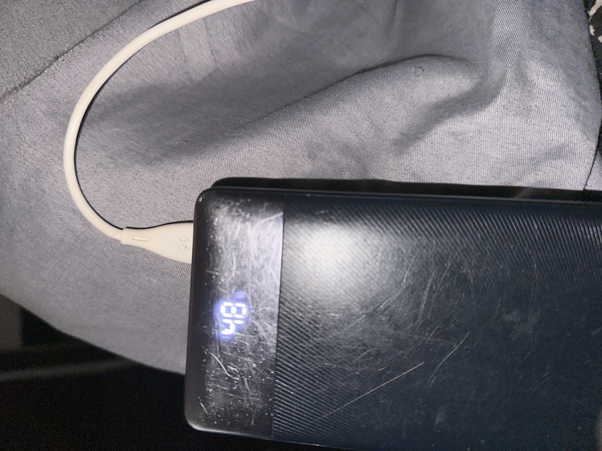 The image shows a black power bank with a digital display showing the number "48", which is likely indicating the battery percentage. A white USB cable is plugged into the power bank. The power bank and cable are resting on a textured gray fabric, possibly clothing. The power bank's surface appears to be scratched. The background is dark and out of focus. There is no discernible indication of the location in Jos, Nigeria.