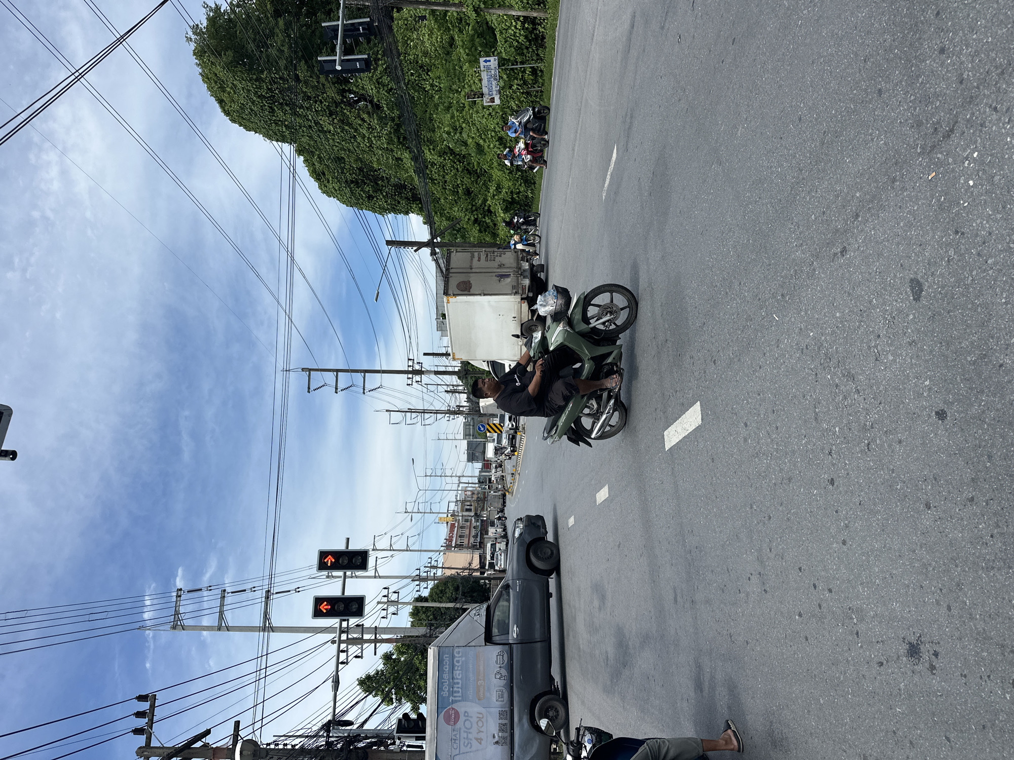 In a medium shot, the image captures a street intersection under a bright, partly cloudy sky. Traffic signals display red arrows, indicating a stop. A dark gray pickup truck with a white box body, advertising "SHOP 4 YOU," is positioned at the intersection. To the right, a man on a green scooter rides through the intersection, wearing a black t-shirt and dark shorts. In the background, a large white truck and several other vehicles are visible, along with dense green foliage and utility poles adorned with numerous wires. The road surface is asphalt, marked with white lines.