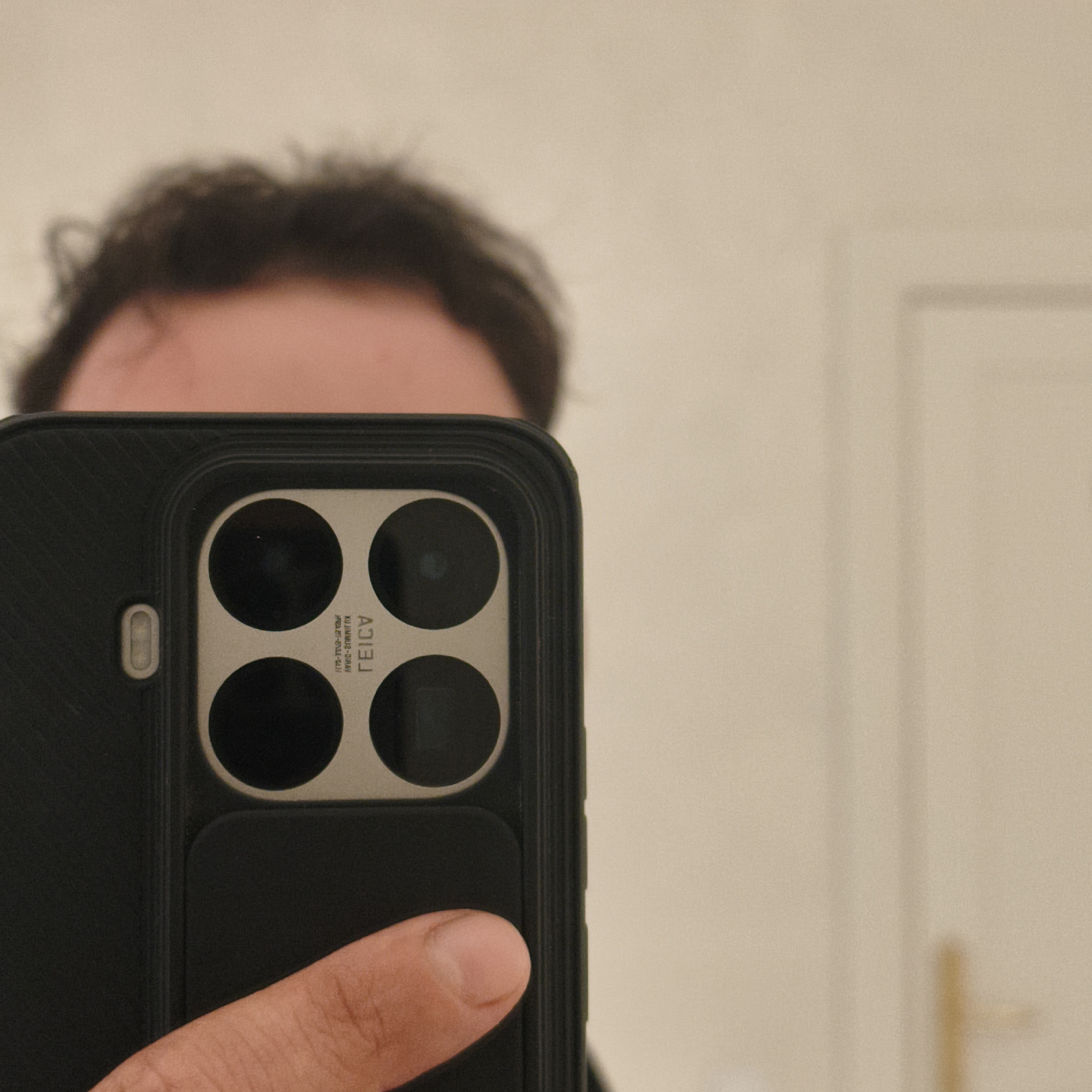 The image shows a close-up of a smartphone held by a person, taken via its rear camera. The phone has a distinctive quad-camera setup housed in a square module. The cameras appear to be circular, with some visible text below them. The phone is in a black case with a textured surface. A finger is visible at the bottom of the frame, holding the phone. The background is out of focus, showing a light-colored wall and what appears to be a door frame. The lighting is soft. The image does not contain any identifiable geographical landmarks or elements that would allow for the determination of its location in Сапоцкінскі сельскі Савет, Belarus.