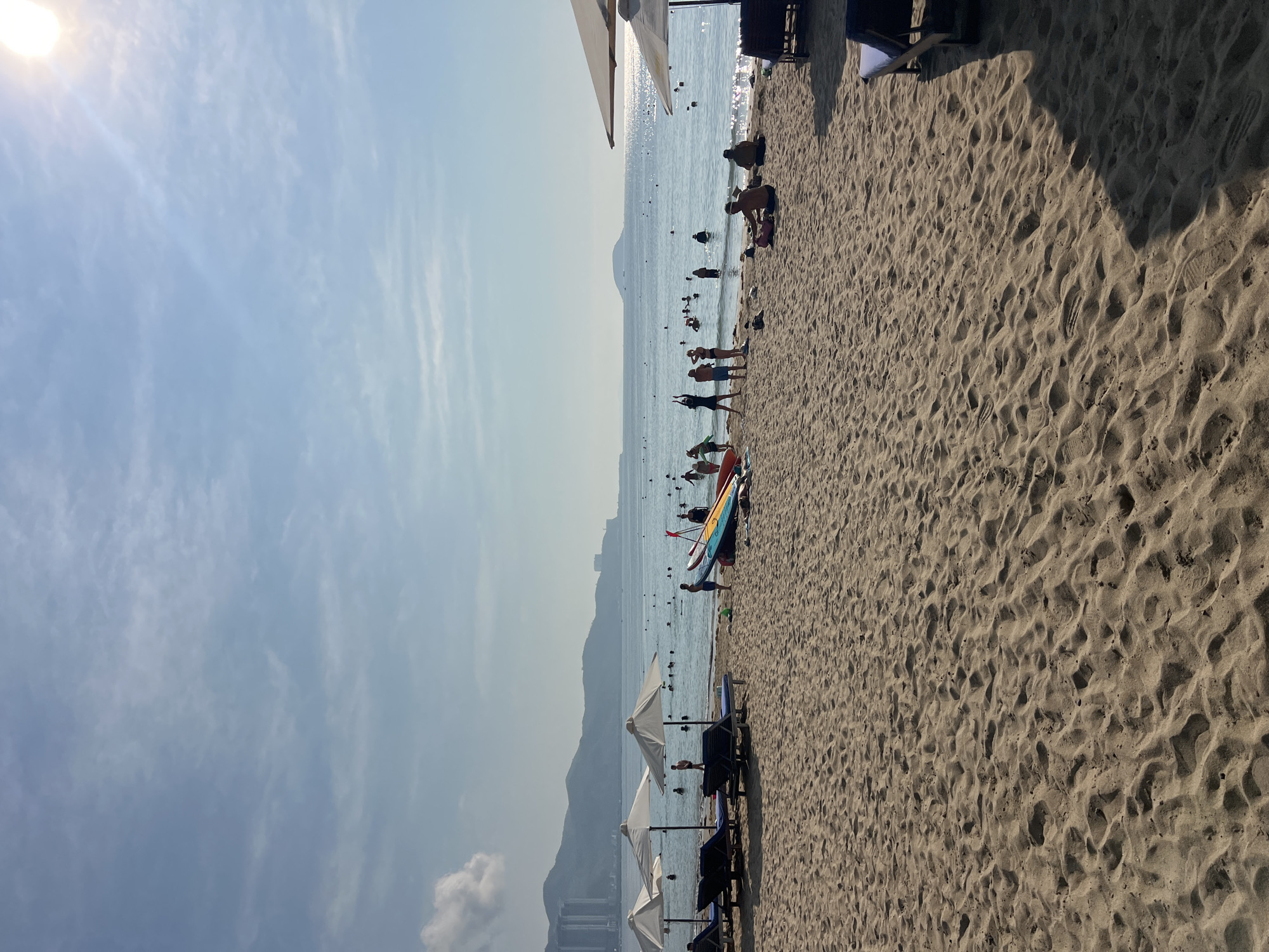 The image depicts a beach scene in Bắc Nha Trang, Vietnam, on a sunny day. The beach is sandy, with clear footprints visible. People are scattered along the shoreline and in the water. Some are swimming, while others are standing or walking. A row of beach umbrellas and lounge chairs are set up on the sand. Surfboards are placed nearby. The ocean stretches out to the horizon, where there is a mountainous shoreline. The sky is blue with scattered clouds.