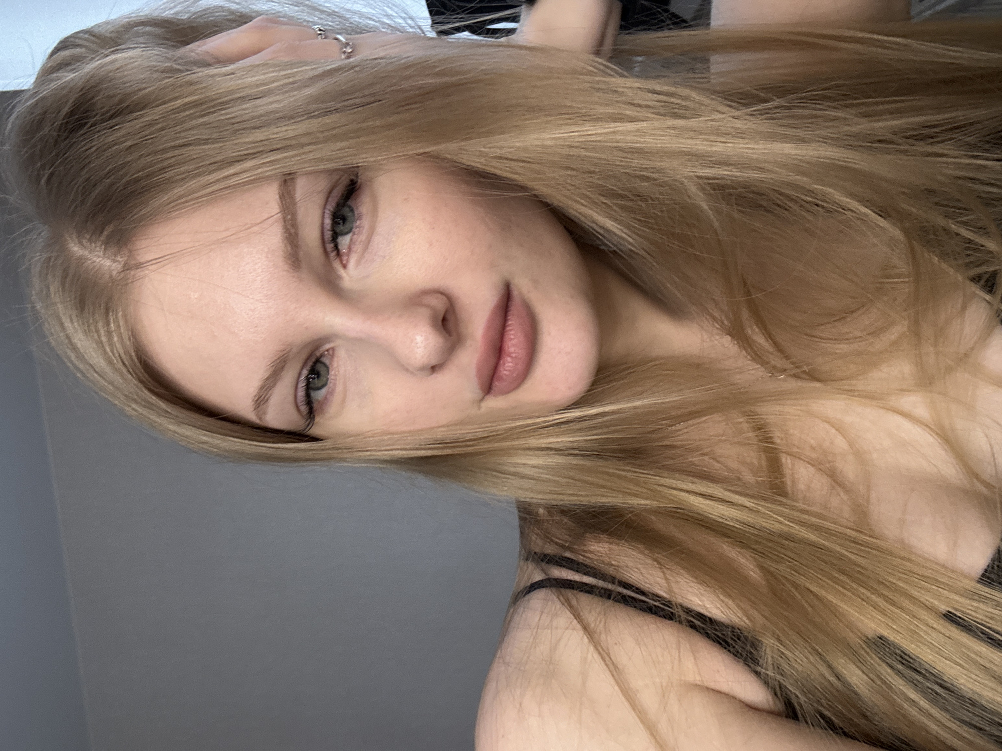 The image displays a close-up, vertically oriented portrait of a young woman, likely in her late teens or early twenties. Her head is tilted, and her long, straight to slightly wavy light blonde or ash blonde hair cascades down, occupying a large portion of the right side of the frame.
The woman has a fair complexion, light-colored eyes (appearing green or hazel) that look directly at the viewer, and subtly applied makeup, including defined eyebrows and light pink or nude lipstick. Her expression is calm and serene, with a slight, subtle smile. Her right hand is visible near her head, with fingers gently touching her hair, and a silver-colored ring is worn on one of her fingers. A black strap, possibly from a top or dress, is visible over her right shoulder.
The background is a plain, solid gray wall, suggesting an indoor setting. The lighting is soft and even, creating a natural and relaxed atmosphere for the portrait. No specific time of day or weather conditions are discernible from the indoor scene. There is no visible text in the image. The provided location context for this image is Perm, Russia.