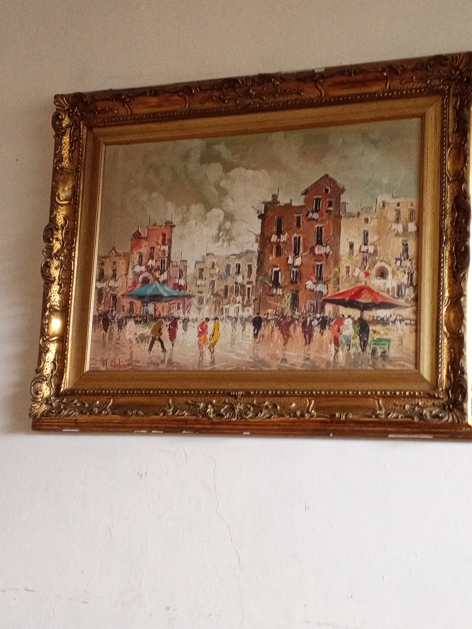 The image depicts a framed painting hanging on a white wall. The painting features a European-style cityscape, possibly a piazza, with buildings, umbrellas, and numerous people. The buildings are multi-story, with terracotta and beige tones. Red and blue umbrellas stand out among the indistinct figures on the ground. The frame is ornate and gilded. The wall beneath the painting is plain white with minor imperfections.