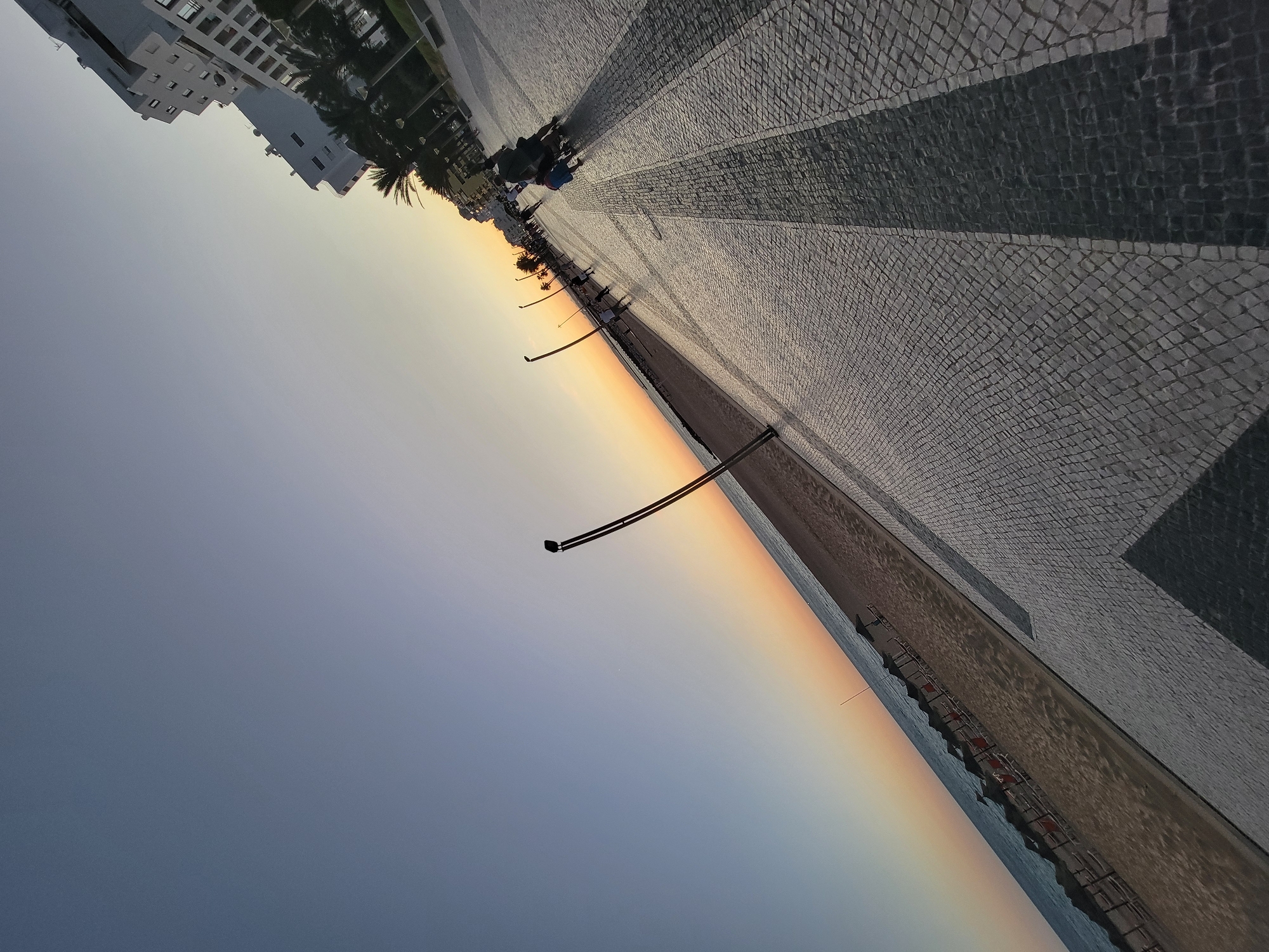 This image depicts a coastal promenade in Quarteira, Portugal, during what appears to be sunset or early evening. The perspective is a low-angle, tilted shot, with the sky occupying the upper left portion of the frame and the ground dominating the right and bottom. The sky exhibits a gradient of colors, transitioning from a deep blue at the top to soft oranges and yellows near the horizon.
To the left, a sandy beach is visible, lined with rows of empty beach chairs and umbrellas. The sea is calm, with gentle waves lapping at the shore. A thin, dark pole stands vertically in the sand, its purpose unclear.
The promenade itself is paved with mosaic tiles, creating a textured surface. Several street lamps with a distinctive, modern design are spaced along the promenade. In the distance, buildings, some appearing to be hotels or apartment complexes, line the edge of the promenade, with palm trees interspersed between them.
Several people are present on the promenade. In the mid-ground, a few individuals are walking, some with strollers. One group appears to be pushing a stroller, and another individual is walking alone. The figures are silhouetted against the brighter sky and illuminated promenade, and their features are not clearly discernible. The overall scene suggests a tranquil and pleasant atmosphere, characteristic of a seaside resort town at dusk.