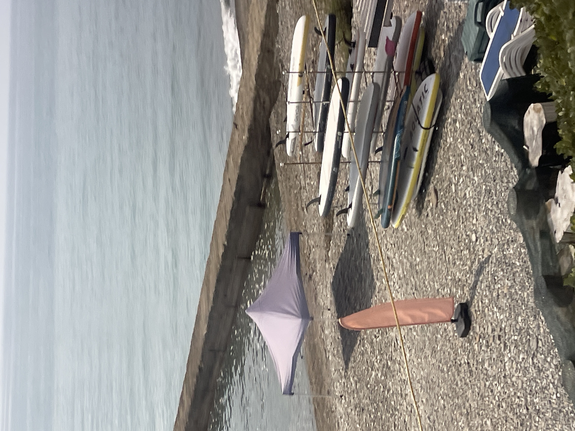 The image depicts a seaside location in Sochi, Russia, on a pebbled beach. In the foreground, a pale orange flag on a stand is visible. Further back, several paddleboards are neatly arranged on racks. A light purple canopy tent is set up on the beach, casting a shadow. Beyond the beach, the calm, gray sea stretches to the horizon under an overcast sky. A concrete seawall runs along the edge of the water. In the lower right corner, some lounge chairs and a green storage box are partially visible, suggesting recreational facilities. The scene appears quiet and possibly unoccupied at the moment.