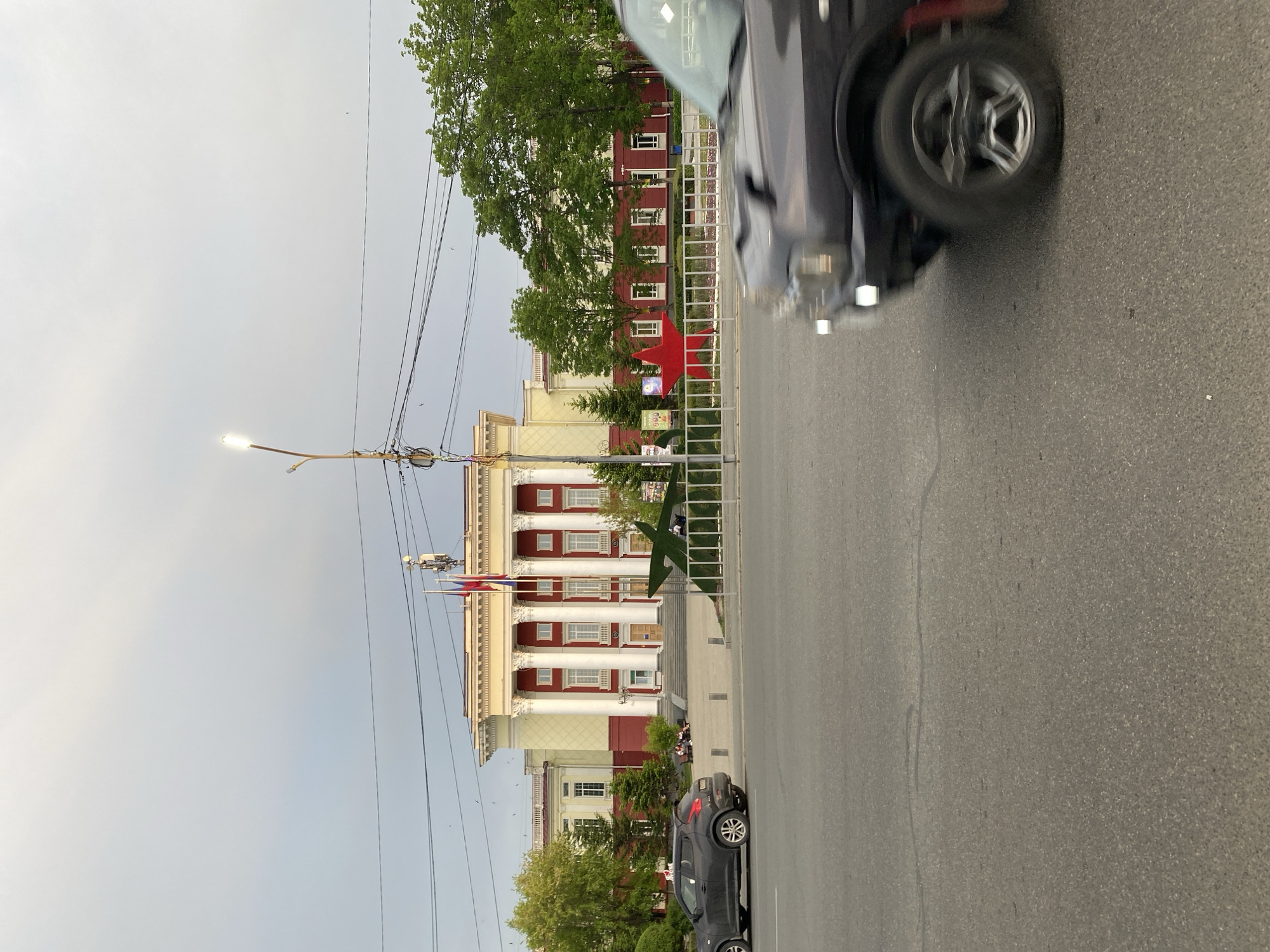 The image captures a view of the city of Artyom, Russia. Visible is a building with a classic facade, featuring columns and a red and yellow color scheme. In front of the building, along the street, a white fence stretches across, adorned with a red star-shaped symbol. An intersection or street runs perpendicular to the viewer's perspective. Overhead, power lines are strung across the sky. In the foreground, on the right side of the frame, is a car in motion. Another vehicle is parked closer to the building.