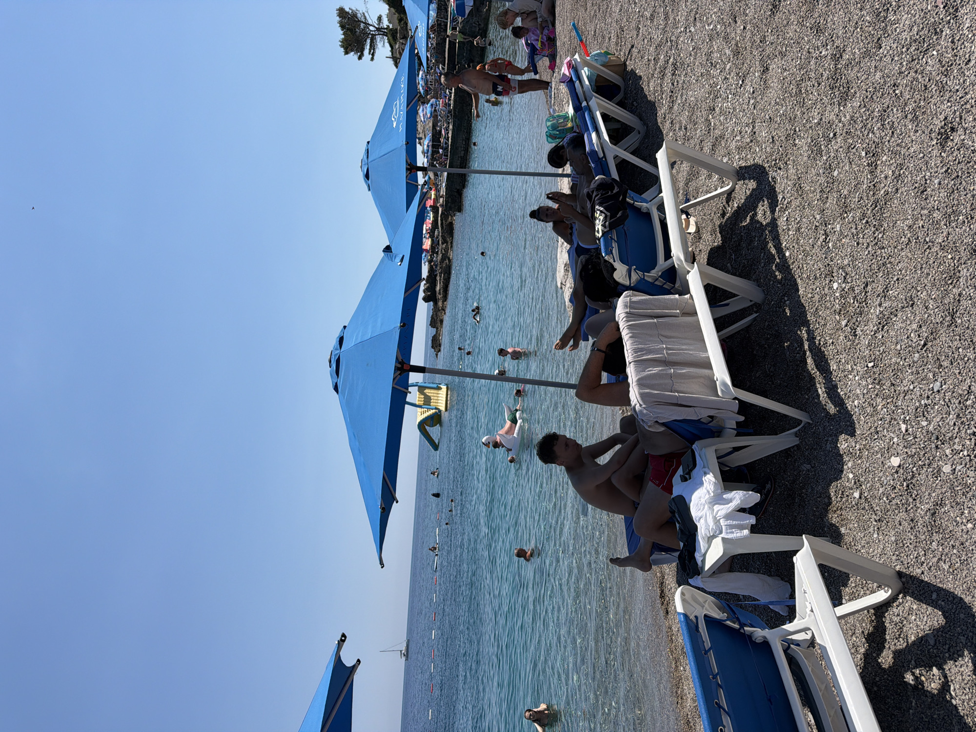 The image depicts a sunny day at a pebble beach in Prijevor, Montenegro. Several blue beach umbrellas are set up, providing shade for lounge chairs on the shore. A group of people are visible relaxing on the lounge chairs, some appear to be reading or using their phones. The sea is clear and blue, with a few individuals swimming or wading near the shore. In the water, a yellow inflatable slide and a large white inflatable swan are present, suggesting recreational activities. Further out in the sea, a sailboat is visible. The beach is covered in small grey pebbles, and the surrounding area features some rocky outcrops and sparse vegetation. The sky is clear and blue.