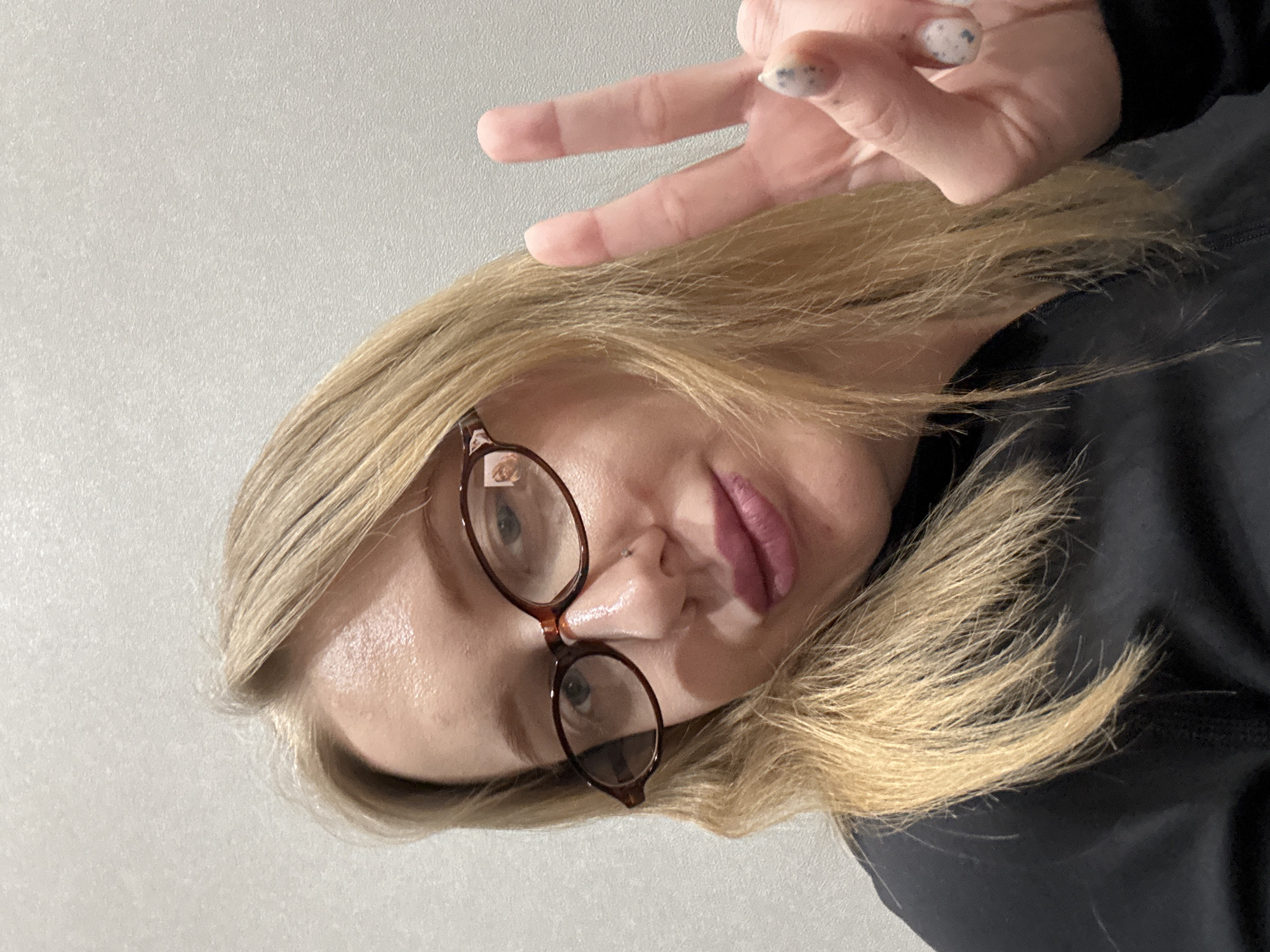 A person with blonde hair, wearing glasses, is holding up two fingers in a peace sign. They are wearing a black garment. The background is a plain, light-colored wall. The location is not discernible from the image.