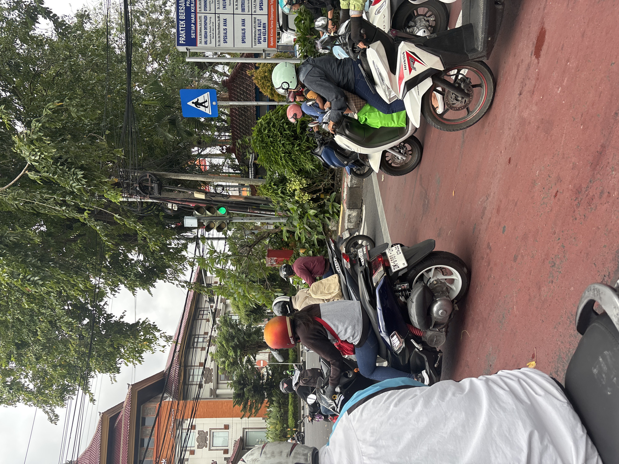 In Denpasar, Indonesia, a busy street scene unfolds with multiple individuals riding motorcycles. The road is paved in red, suggesting it may be a pedestrian or dedicated motorcycle lane. Traffic lights, currently displaying green, indicate a flow of movement. A prominent blue sign with a white pedestrian silhouette signifies a crosswalk. Buildings with traditional Balinese architecture, featuring intricate stone carvings and tiled roofs, line the background. Lush green trees provide a natural canopy over the urban environment. People, predominantly wearing helmets for safety, are engaged in the act of riding their scooters. One individual in the foreground, wearing a white shirt, faces away from the camera, while others are captured in profile or from behind as they navigate the street. The overall impression is one of daily life in a bustling Indonesian city, where motorbikes are a primary mode of transportation.