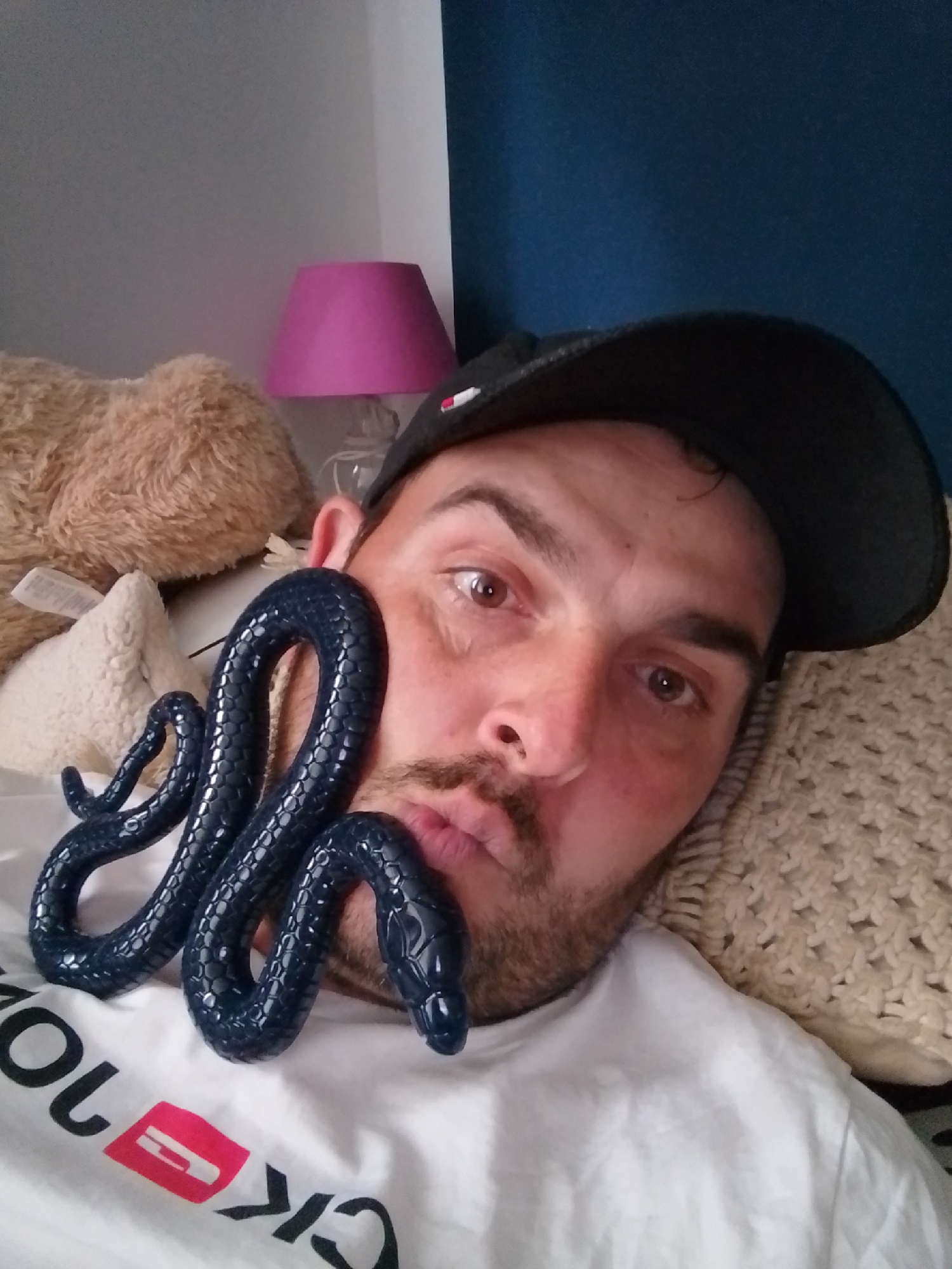A man, wearing a black baseball cap and a white t-shirt with a red and white logo, lies on a bed. He is looking at the camera and has a dark blue toy snake wrapped around his face and neck. The snake appears to be made of a glossy material. Behind him, a beige teddy bear and a white textured cushion are visible. A pink lampshade is in the background on the left. The overall setting suggests an indoor environment, possibly a bedroom.