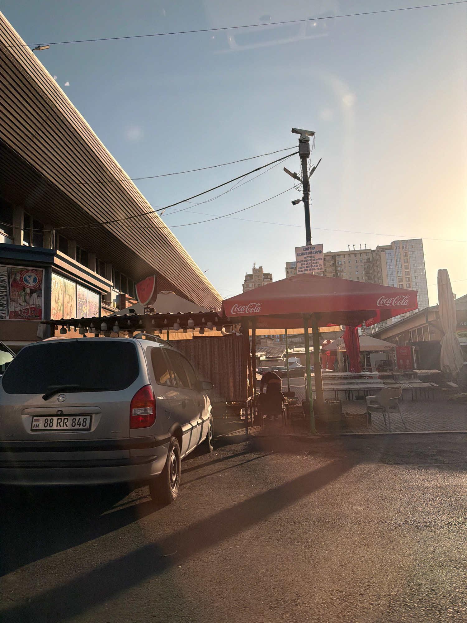 The image captures a street scene in Yerevan, Armenia, likely during the golden hour due to the warm, low-angled sunlight casting long shadows. In the foreground, a silver Opel Zafira wagon with license plate "88 RR 848" is parked on an asphalt road. To the left, a modern building with a sloped, ribbed facade and large windows is visible. A red Coca-Cola umbrella shades an outdoor seating area with tables and chairs. Behind this, a street light pole with attached cameras and electrical wires stands tall. Further in the background, several residential buildings are seen against the bright, sunlit sky. A person is seated under the umbrella, seemingly engaged with something on a table. The overall impression is one of an urban marketplace or a casual dining spot.
