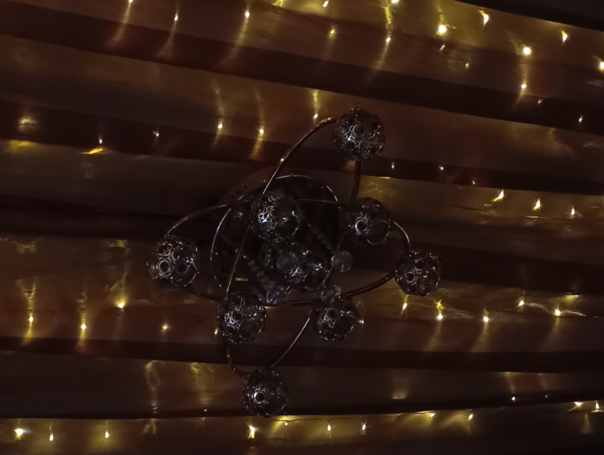 The image depicts a decorative lighting fixture, possibly a chandelier, with several spherical elements featuring intricate cut-out designs. The fixture is metallic, with thin, curving arms extending from a central point to support the spheres. The background appears to be a series of horizontal, light-colored, possibly wooden, panels or boards, illuminated by strings of small, warm-toned lights. The scene suggests an interior setting with ambient lighting. There is no evidence to support any mention of Krasnodar, Russia.
