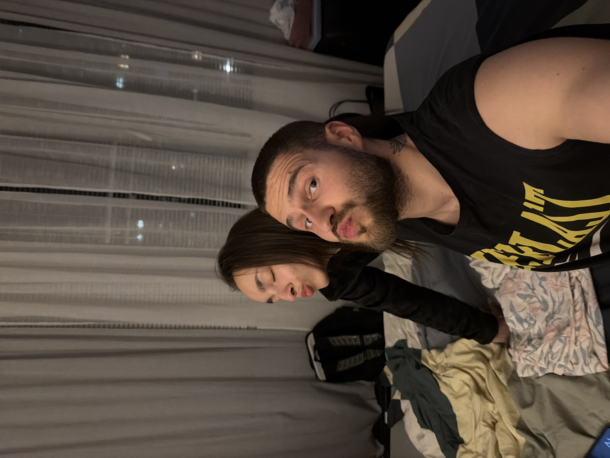 An intimate indoor scene captures two individuals, a man and a woman, posing closely together, likely for a selfie. The image is taken at night, with reflections of city lights visible through horizontal blinds or sheer curtains in the background, suggesting an urban setting in Sofia, Bulgaria.
The man, with dark hair and a beard, is wearing a black t-shirt with a yellow graphic design on the front. He is looking towards the camera with a playful puckered, kissing expression. A tattoo is visible on his neck. The woman, with long dark hair and wearing a dark long-sleeved top, leans her head against the man's. Her eyes are closed, and her lips are also pursed in a matching kissing expression.
They appear to be on or near a bed, which is covered with various fabrics: a plain grey sheet, a light-colored floral patterned fabric with pink and blue elements, and crumpled solid-colored fabrics in shades of beige/yellow and dark green. A black bag is partially visible behind the woman. The overall lighting is dim, contributing to the cozy and personal atmosphere. No clearly legible text is visible on clothing or other objects.