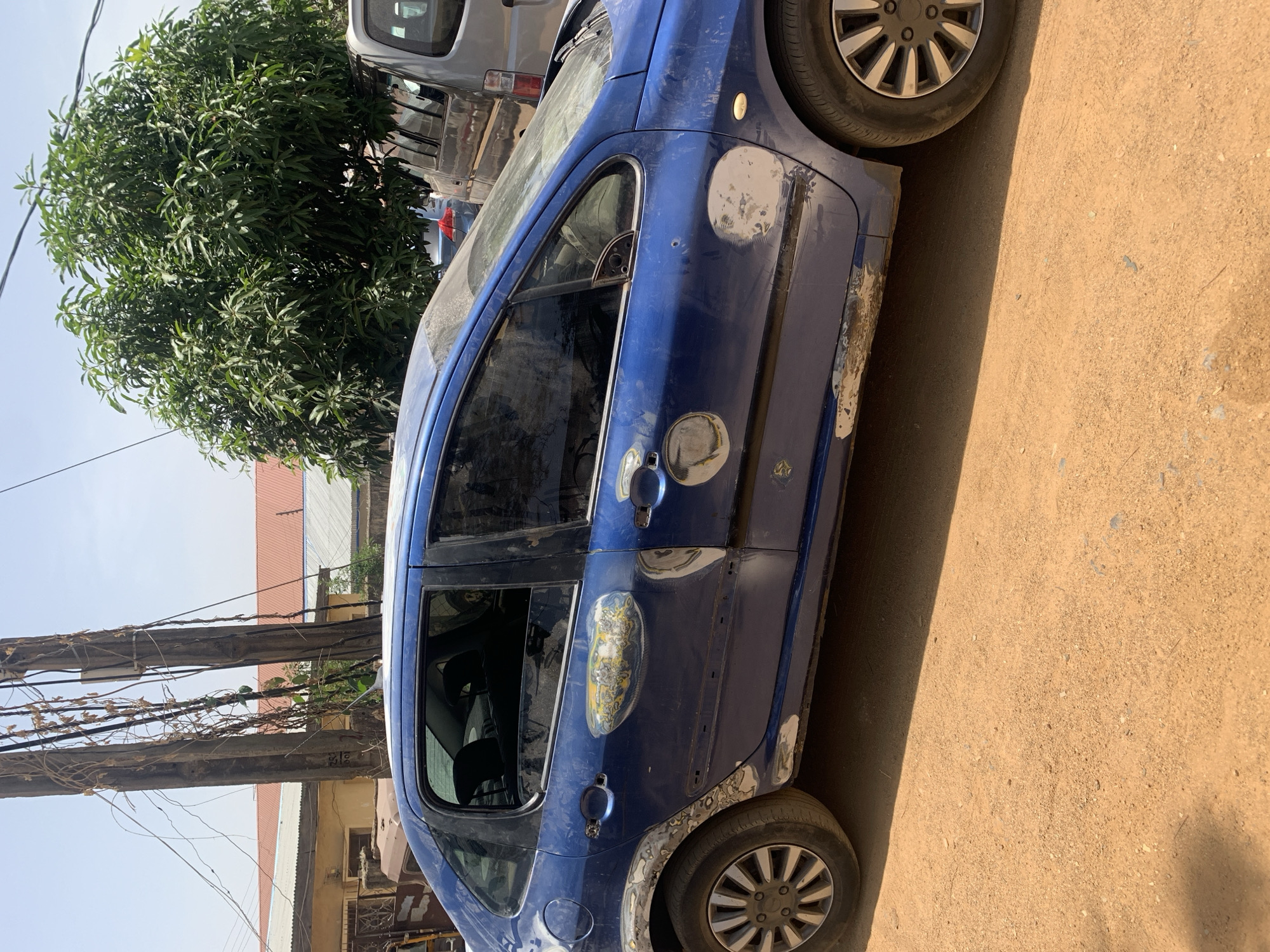 A blue four-door hatchback car is parked on a dirt road in Rafin Par, Nigeria. The car appears to be damaged, with body filler patches and missing paint. A tree is visible in the background, along with utility poles and buildings. Another vehicle is parked behind the blue car.