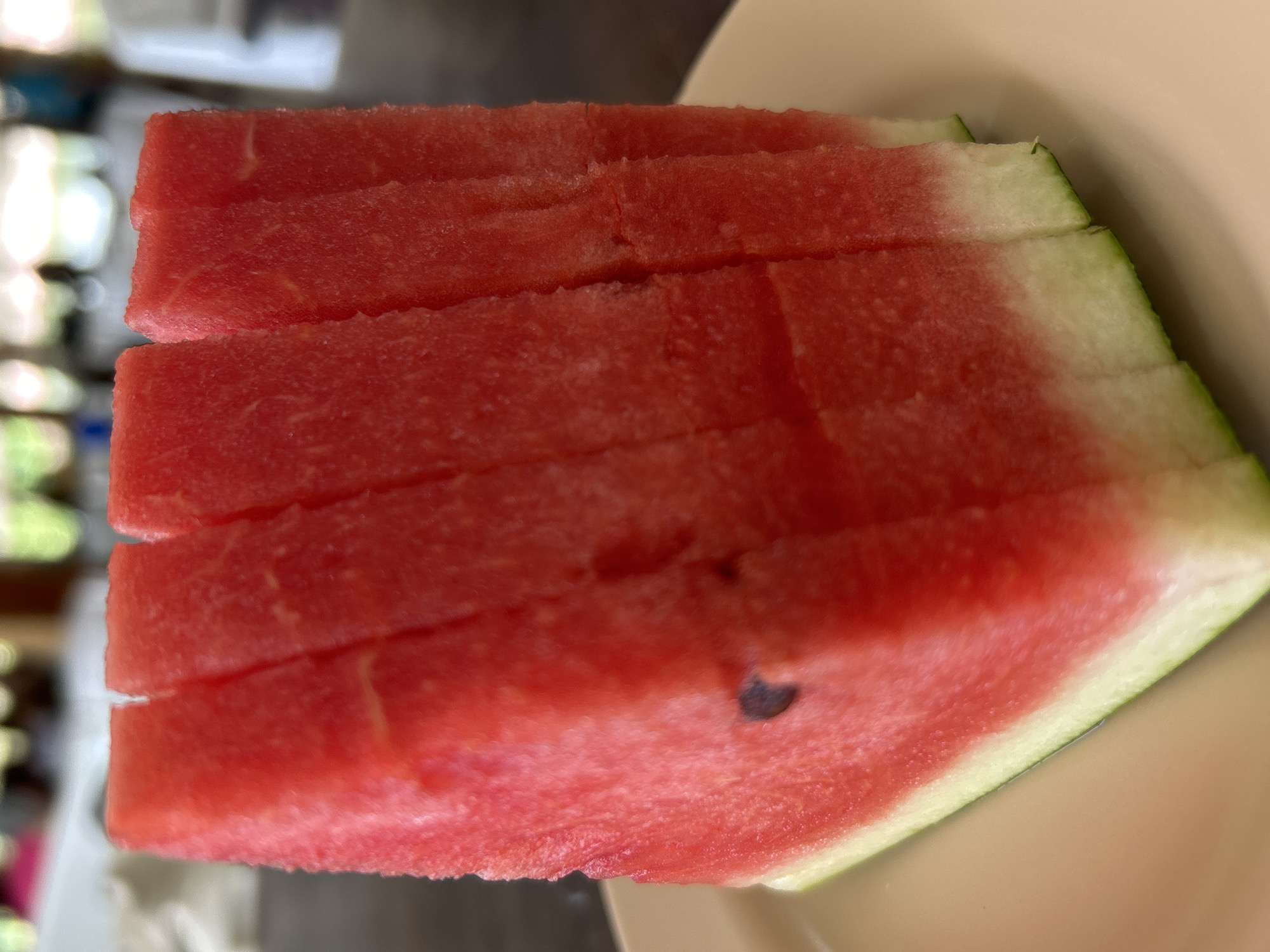 The image displays a close-up of multiple slices of red watermelon, likely cut into thick wedges or rectangular pieces, arranged on a light beige or cream-colored plate. The watermelon flesh is a vibrant crimson, exhibiting a slightly granular texture and faint, lighter red or white striations, indicative of its natural fibrous structure. A small, dark, oblong seed is visible embedded in the leftmost slice. The bottom portion of the slices reveals the green outer rind transitioning to a pale green and then white inner rind, suggesting a complete cut. The background is significantly blurred, featuring indistinct warm and cool tones, hinting at an indoor or shaded outdoor setting with ambient natural light. The out-of-focus elements suggest a casual environment, potentially a dining area, consistent with a resort or restaurant in Ao Nang, Thailand. No individuals, overt actions, or interactions are discernable within the frame.