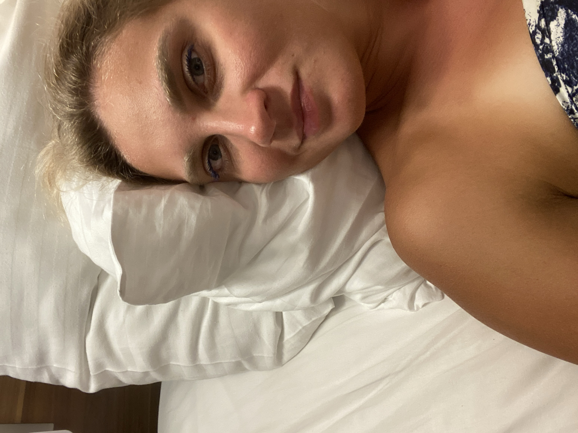 A woman with blonde hair and blue eyeliner lies in a bed, resting on white pillows. She is wearing a strapless top with a blue and white patterned trim. The lighting suggests an indoor setting, possibly a hotel room. The overall scene is casual and relaxed. The location is unknown.