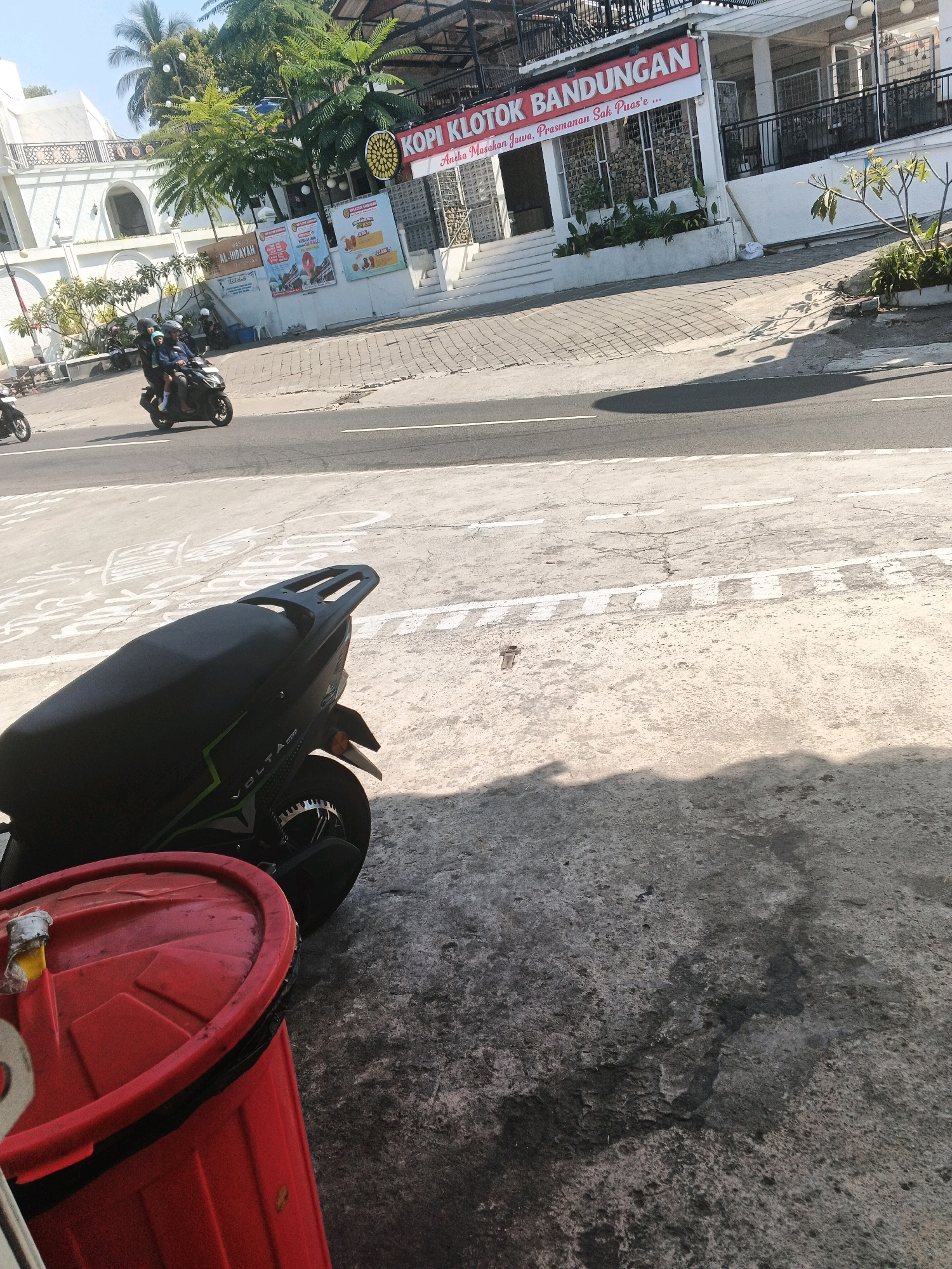 The image captures a street scene in Pakopen, Indonesia, bathed in sunlight. In the foreground, a black motorcycle with green accents is parked on a concrete surface, partially obscuring a bright red trash bin. The motorcycle's rear wheel and part of its seat are visible. Further back, a family of three rides on another motorcycle, a man driving with two passengers. They are on a paved road with white lane markings.
On the right side of the road, a building with a red sign reading "KOPI KLOTOK BANDUNGAN" stands prominently. The building appears to be a restaurant or cafe, with outdoor seating and greenery. A white building with a balcony is visible to the left of the cafe. Several other motorcycles are parked or in motion in the background, suggesting a busy street. The overall environment is urban, with palm trees and other vegetation adding to the tropical atmosphere. The ground is a mix of concrete and asphalt, with some areas showing wear and tear. Shadows are cast across the road and ground, indicating a sunny day.