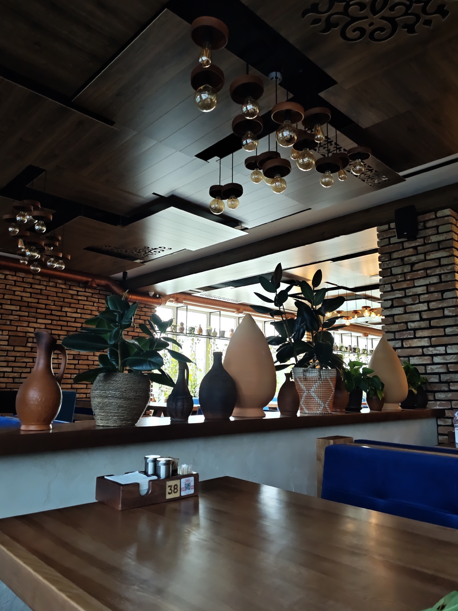 The photograph captures a restaurant interior, likely in Vinnytsia, Ukraine. The focal point is a row of decorative objects and plants atop a low wall or partition. These include ceramic vases and potted rubber plants. Behind this partition, a glimpse of a window shows additional objects and greenery.
The room features a complex ceiling design composed of various wood paneling, some with decorative cutouts, and several clusters of pendant lights. The wall adjacent to the partition is constructed of red bricks.
In the foreground, a wooden table is visible, with a table number (38) and condiment holder placed on it. Part of a blue banquette is also visible, suggesting seating for patrons. The setting appears to be a stylish or upscale establishment. There are no people visible in the image.