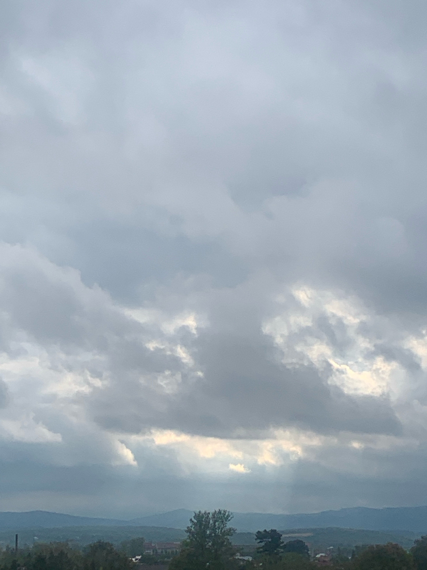 This is a photograph of a cloudy sky over a distant landscape. The sky is filled with a mix of dark and light gray clouds, with some patches of brighter light peeking through. Below the clouds, rolling hills and mountains are visible in the distance. The foreground shows a dark silhouette of trees and some buildings, suggesting a rural or suburban setting. The overall mood of the image is somewhat overcast and moody. The location is identified as the city of Kolomyia, Ukraine. There are no people, subjects, or actions depicted in the image.