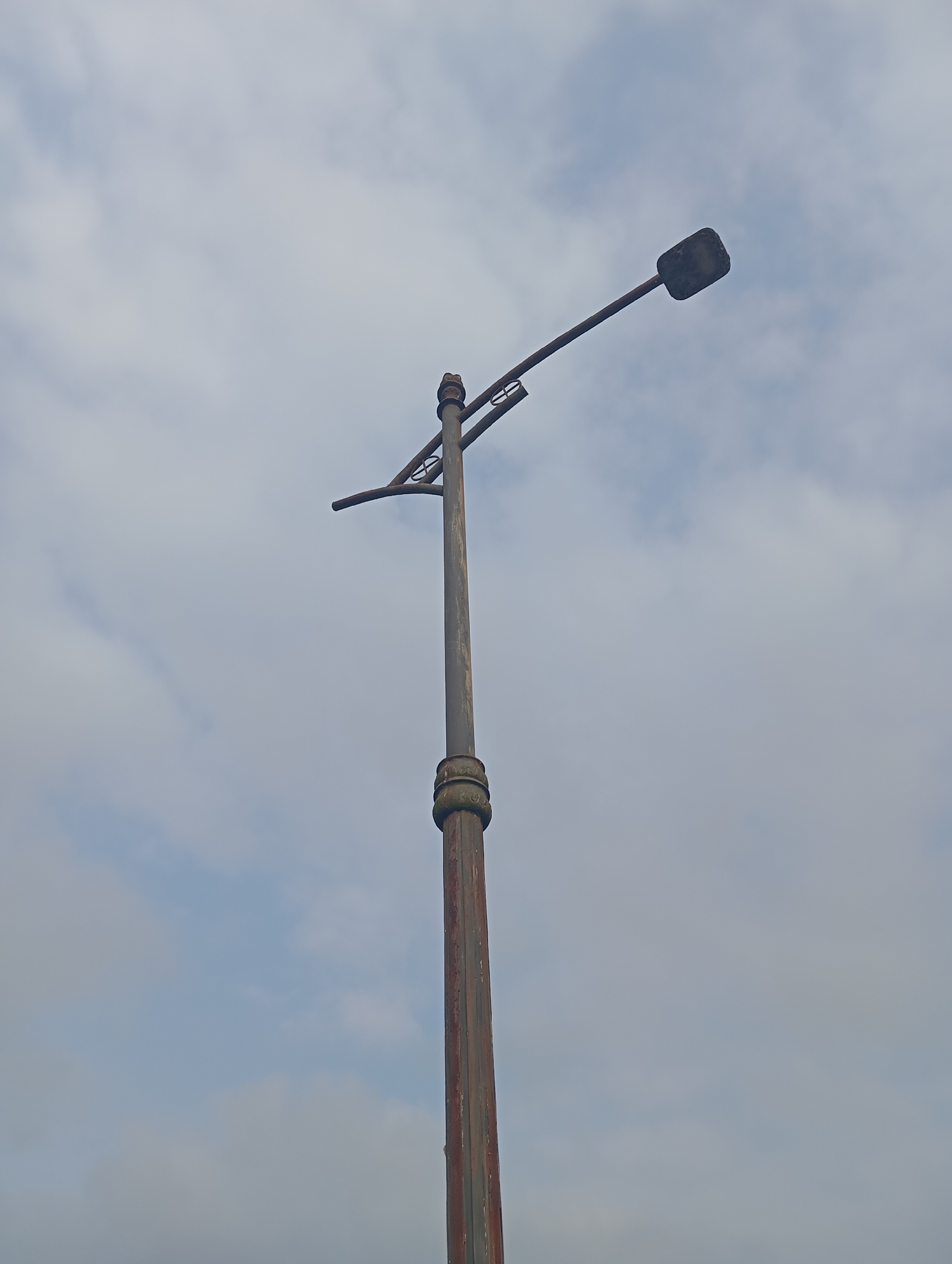 The image shows a street lamp against a cloudy sky. The lamp is made of metal, with a single light fixture at the end of an angled arm. The pole of the lamp is cylindrical and appears to be weathered or rusted in places. There are decorative bands around the top and bottom of the main section of the pole. The lamp is positioned in the lower right quadrant of the image, extending upwards and to the left. The background is entirely composed of a sky with scattered clouds, appearing overcast. The scene is devoid of any people, animals, vehicles, or other urban infrastructure, focusing solely on the lamp and the sky. The setting of Kedungbenda, Indonesia, is not visually discernible from the image itself.