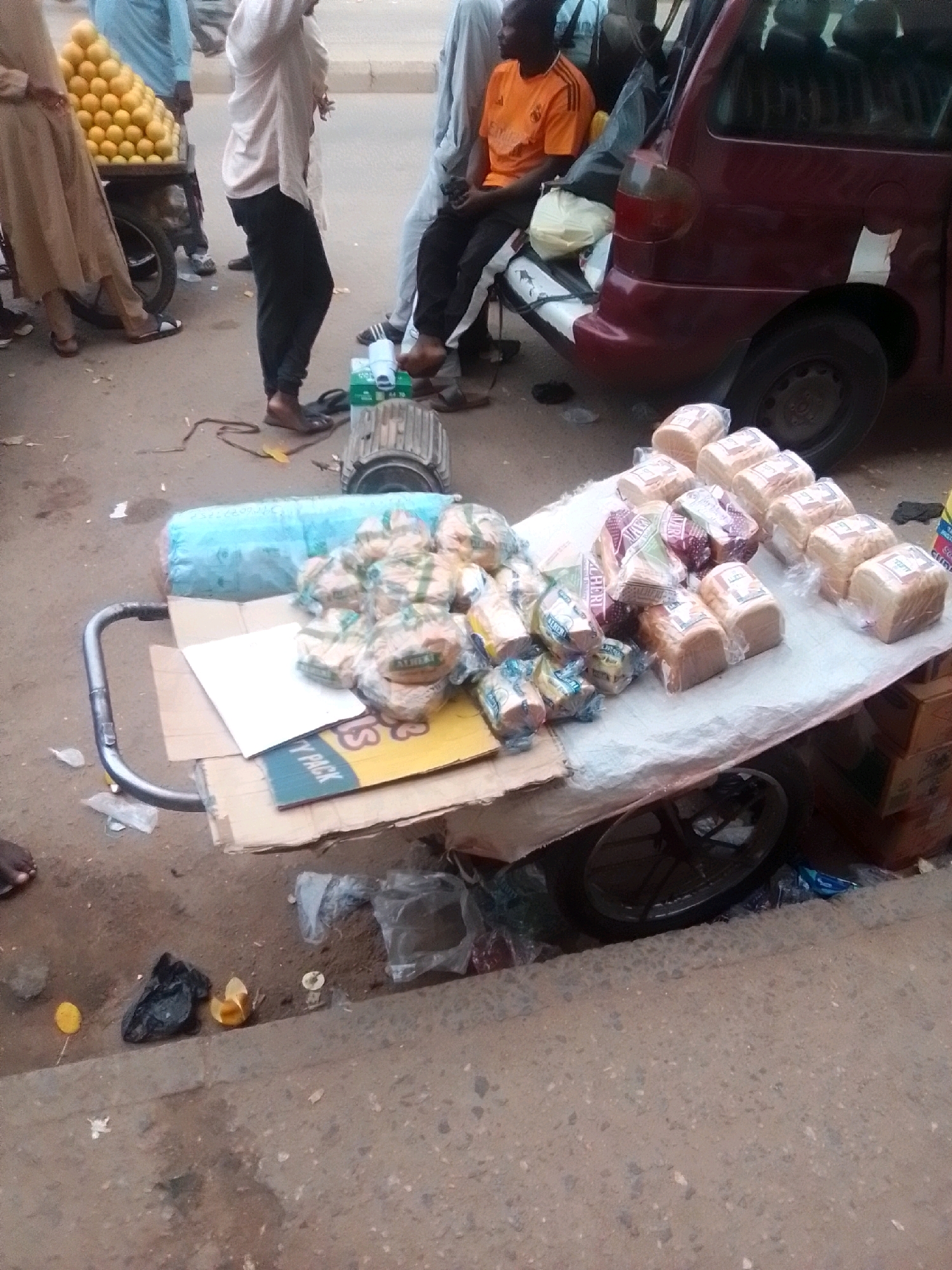 The image depicts an outdoor scene, possibly a street market, in Arawa, Nigeria. In the foreground, a cart with a white tablecloth is laden with many loaves of bread packaged in clear plastic. The loaves are of various sizes and some have visible branding. To the left of the cart, a large blue plastic-wrapped item is visible. A metal object, possibly an engine or generator, sits on the ground behind the cart. In the background, several people are present. A person in an orange shirt is seated near a burgundy van. Another person in a light-colored outfit is standing next to a cart piled high with yellow fruits, likely oranges. The ground is a dirt or paved surface with scattered debris, including plastic bags and wrappers. The overall impression is that of a bustling street vending area.