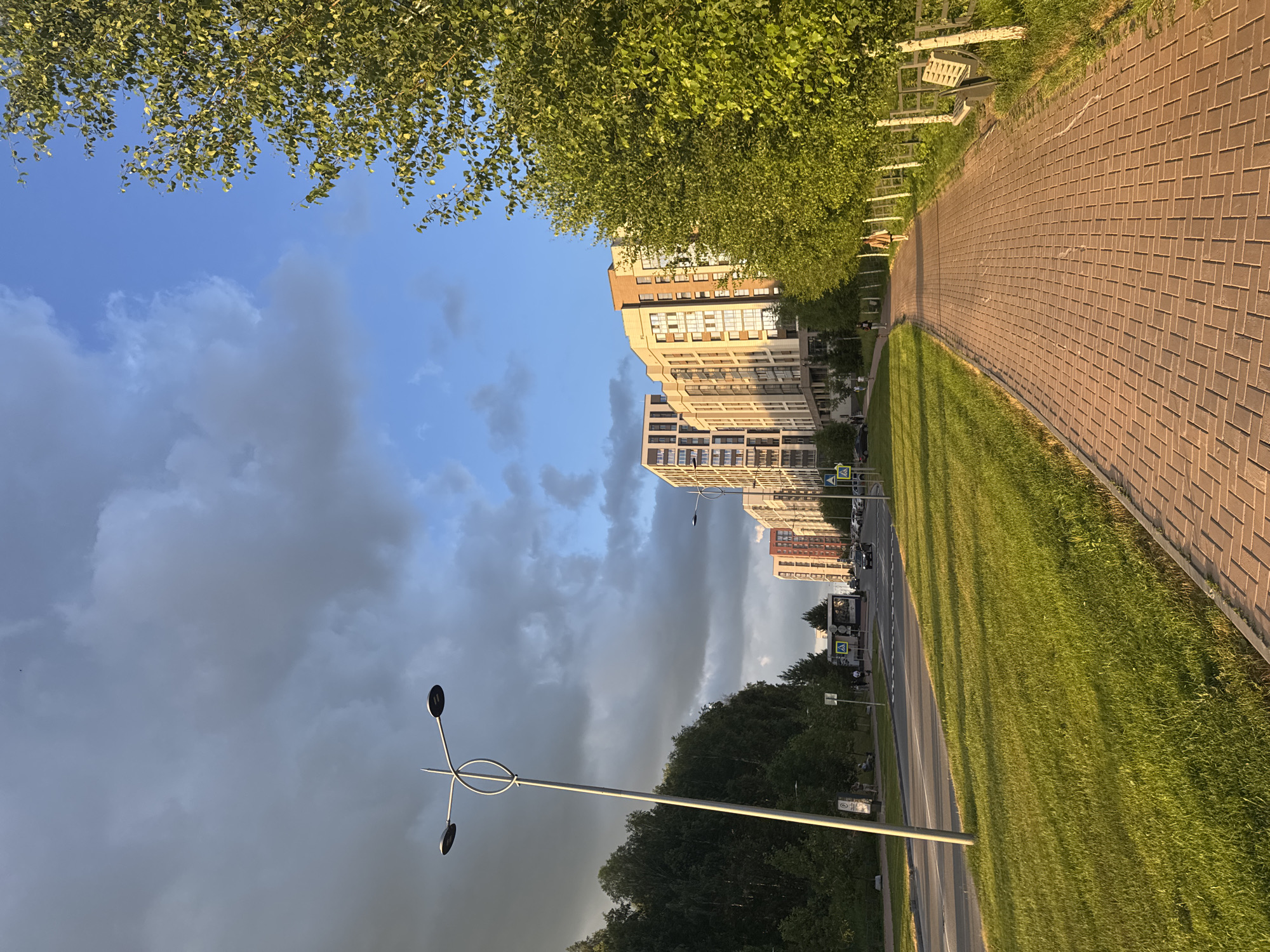 This image depicts a street scene in Koltsovo, Russia, likely during the late afternoon or early evening, as indicated by the warm, golden light casting long shadows across the meticulously paved walkway and the lush green grass. The scene is dominated by a row of modern, multi-story apartment buildings that are predominantly beige and light brown, with numerous windows reflecting the sky. The buildings appear residential and well-maintained.
To the left of the buildings, a vibrant green canopy of trees, possibly birch trees due to their leaf shape and density, overhangs the scene. The sky above is a mix of bright blue and dramatic, dark gray clouds, suggesting a change in weather or the lingering presence of a storm. A modern street lamp with a unique, sculptural design stands tall in the foreground, its metallic structure catching the light.
The paved walkway curves alongside the road, bordered by well-kept grass. The road itself is visible, with a few cars parked or driving along it, and there are pedestrian crossings and street signs, including a blue sign with a yellow pedestrian symbol. A bus stop shelter is also visible. While there are a few indistinct figures on the walkway and near the bus stop, no specific individuals or activities are clearly discernible. The overall impression is one of a modern, developing urban or suburban environment bathed in the golden light of a late day, under a dynamic sky.