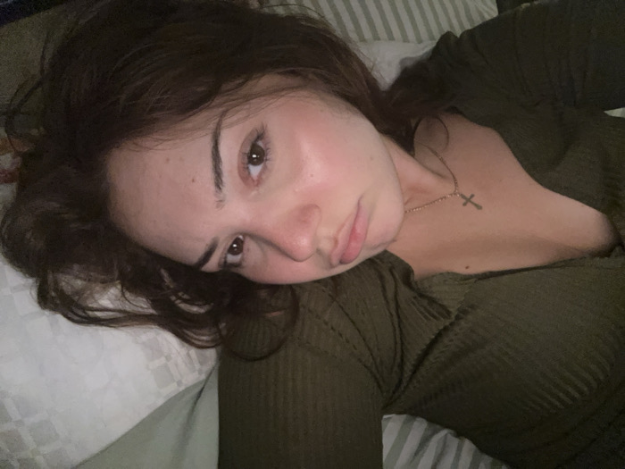 A young woman with dark hair and brown eyes lies on a pillow with a white and gray patterned cover. She wears an olive green, textured top. A gold necklace with a cross pendant is visible. She looks at the camera with a slight pout. The image is dimly lit, creating a soft, intimate atmosphere.