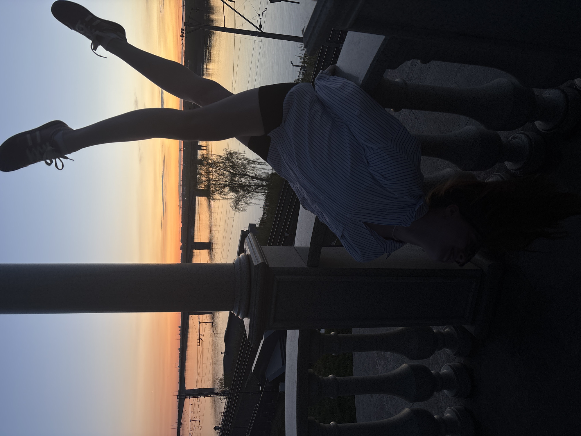 A woman is standing on a stone railing on a bridge overlooking the Kama River in Perm, Russia. She is leaning forward with her hands on the railing and her legs extended upwards at approximately 45-degree angles. She is wearing shoes, black shorts, and a light-colored striped shirt. The sky is gradient of colors from blue to orange, indicating either sunrise or sunset. A bridge and power lines are visible in the background.