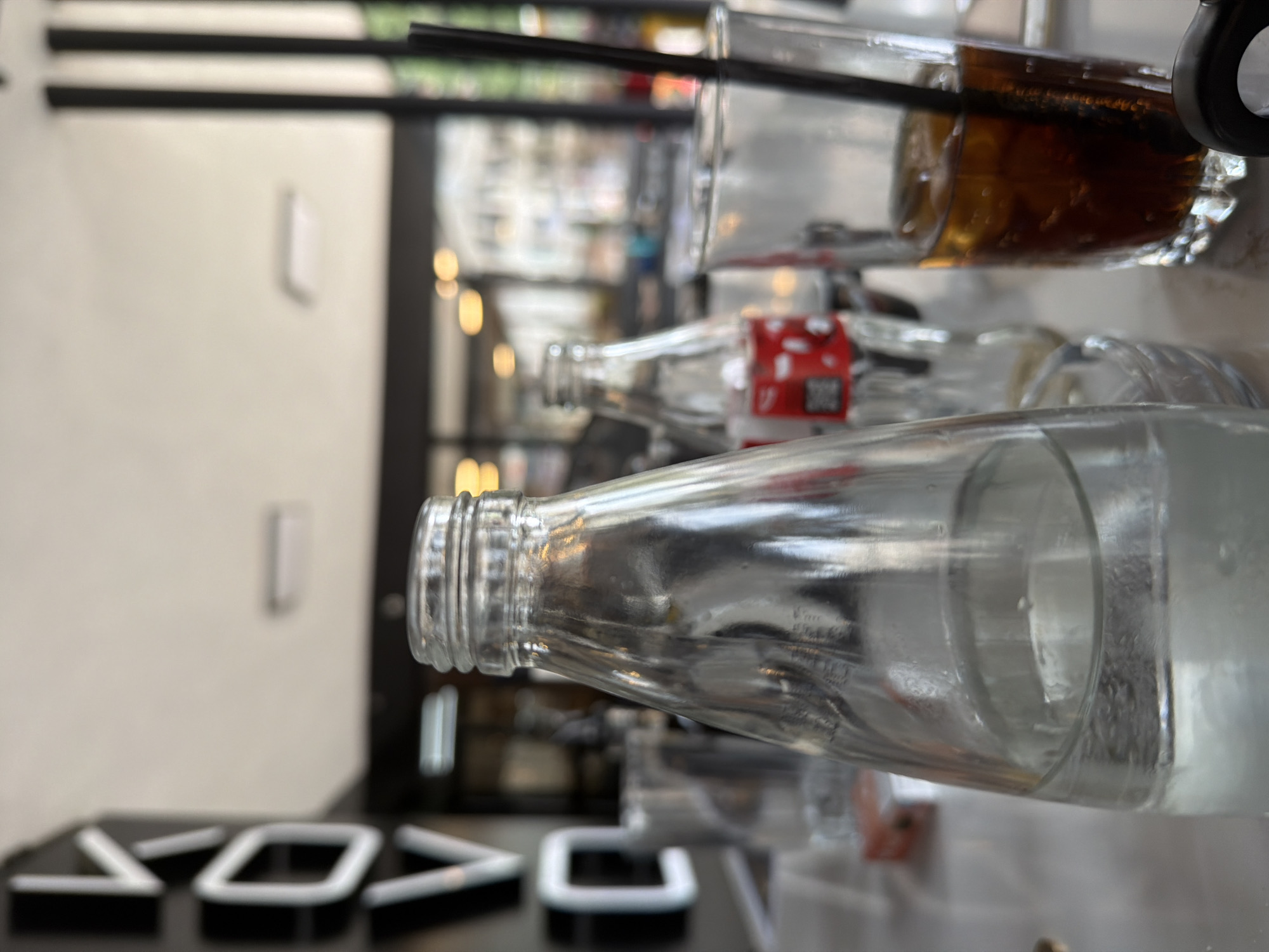The image depicts a close-up, slightly low-angle view of a tabletop scene within a commercial establishment, specifically a cafe or restaurant, located in Etropole, Bulgaria. In the foreground, a clear glass bottle, approximately half-full with still water, is prominently centered. To its right and slightly behind, a second clear glass bottle, partially obscured, displays a red and white Coca-Cola label on its lower portion. Further to the right, a clear glass contains a dark, carbonated beverage with several ice cubes and a black drinking straw. A small, rectangular object, possibly a cigarette pack with red markings, is partially visible on the table surface behind the water bottle. The table itself appears to be a light-colored, possibly marble or laminate, surface.
The background is significantly out of focus, suggesting a shallow depth of field. On the far left, large, stylized white letters on a dark background are visible, forming what appears to be part of a sign, with the letters "KOVO" being discernible from top to bottom. Beyond these elements, blurred indoor lighting fixtures and indistinct architectural features are visible, indicating an interior setting with natural light sources, possibly large windows or an open facade. No individuals are directly visible in the frame.