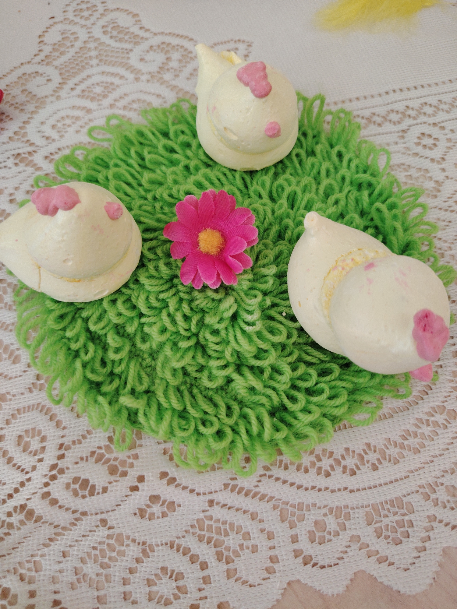 The image depicts a display of three cream-colored, snail-shaped meringues decorated with pink dots and a pink flower. They are arranged on a circular, green, yarn-like mat placed on a white, lacy tablecloth. The location of the image is unknown.